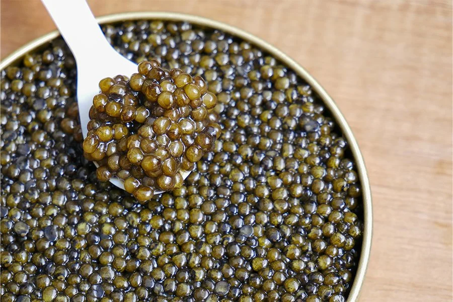 A spoon scooping black caviar from a gold-colored tin container.
