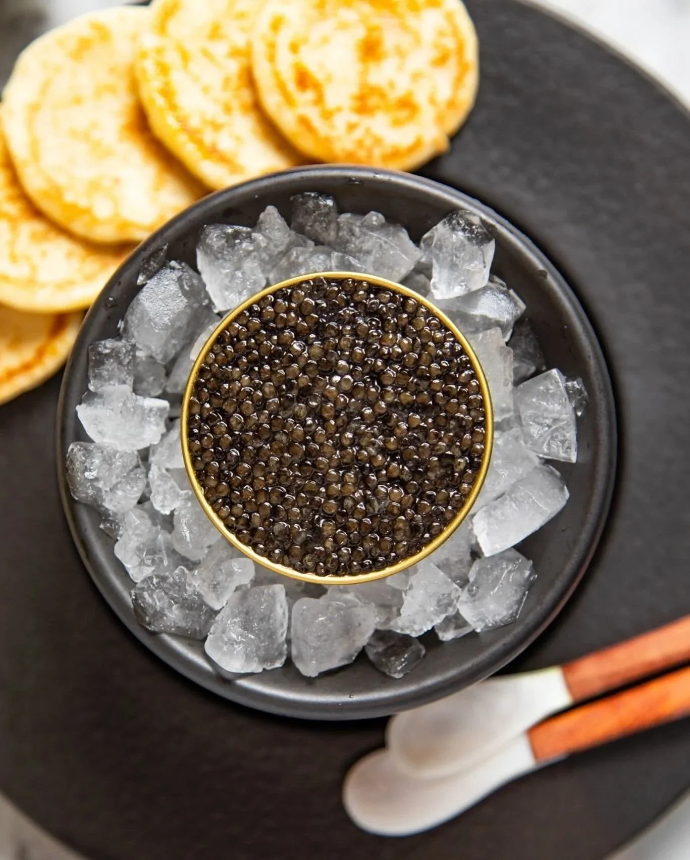 A small bowl of black caviar on ice, surrounded by a larger bowl filled with ice, with sliced bread in the background, on a dark tray.
