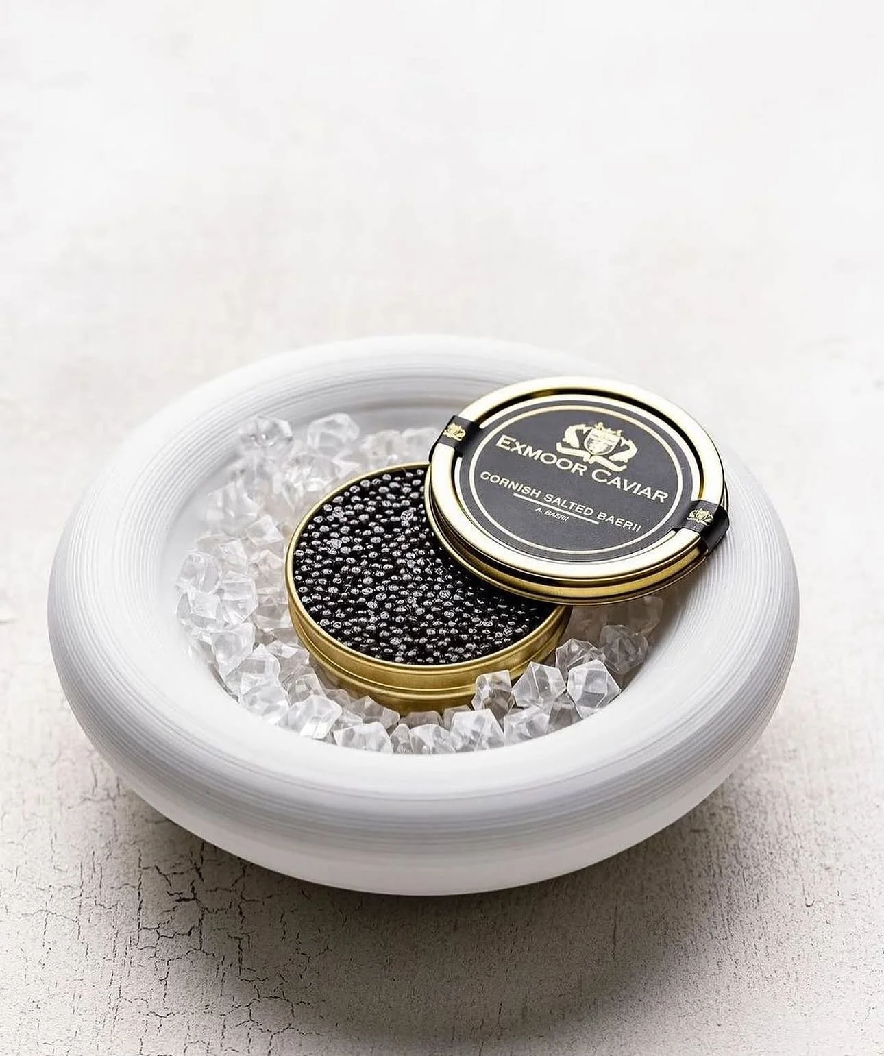 A round white bowl filled with ice and a small metal tin of Exmoor Caviar, with the caviar inside visible.