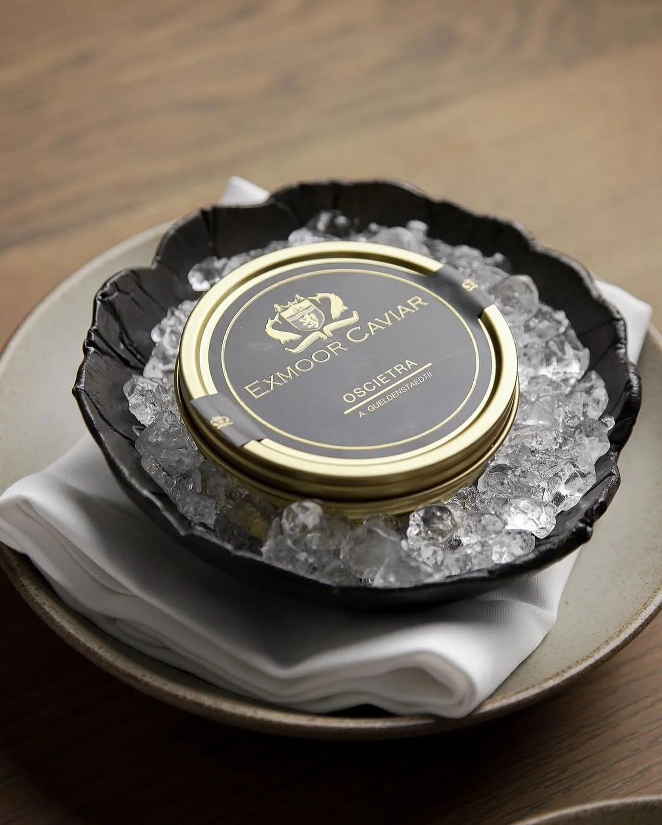 Canned Exmoor Caviar in a black tin with gold accents, resting on ice in a black dish, served on a white napkin on a beige plate.