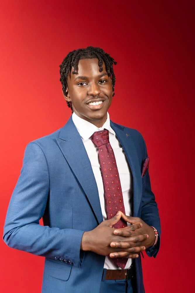 Young man in a blue suit with a white shirt and red patterned tie, smiling and hands clasped in front, against a red background.