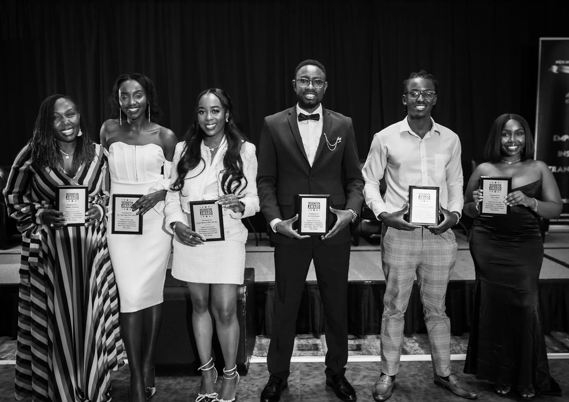 Group of six diverse people standing on stage at an awards ceremony, holding framed awards, dressed in formal attire.