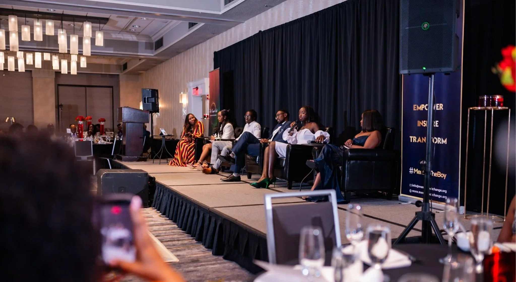 Panel of six diverse speakers seated on stage at a conference, with a black curtain backdrop, a podium, and a banner that says 'Empower, Inspire, Transform'.