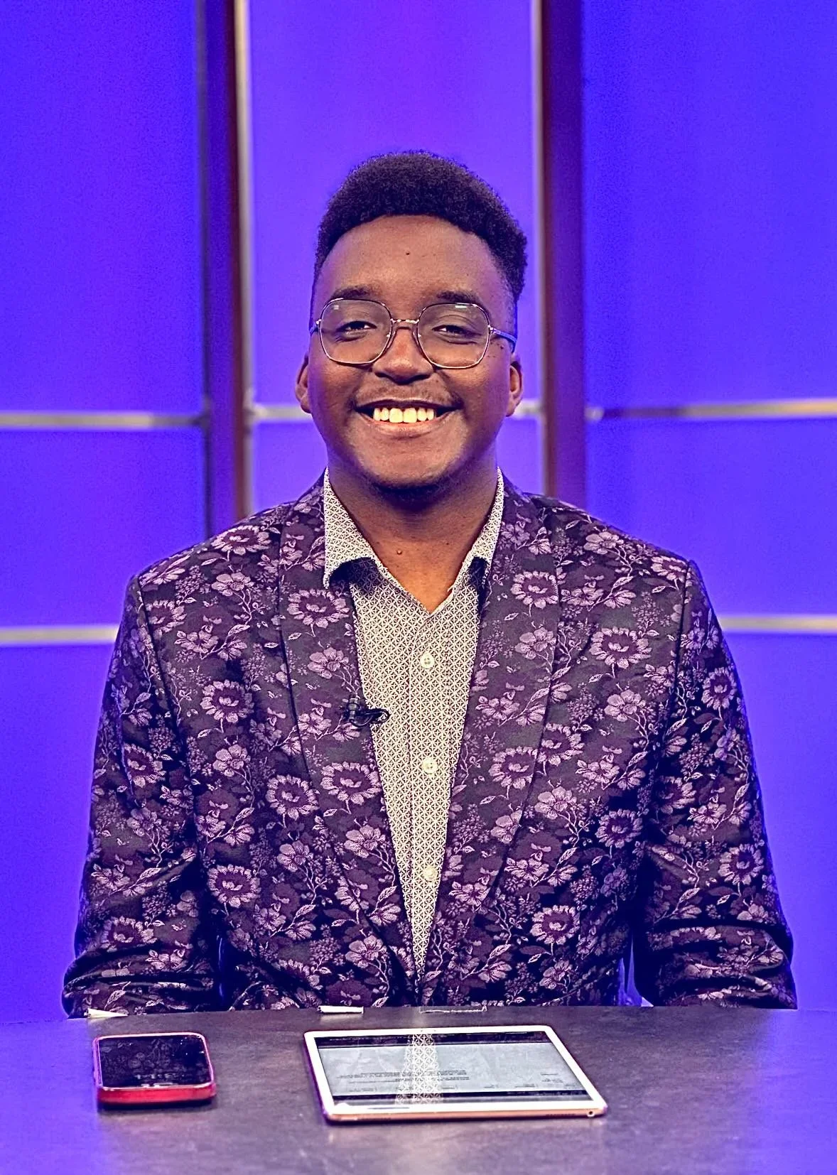 A smiling man sitting at a table with a smartphone and a tablet in front of him, wearing glasses and a floral patterned blazer, against a purple background.