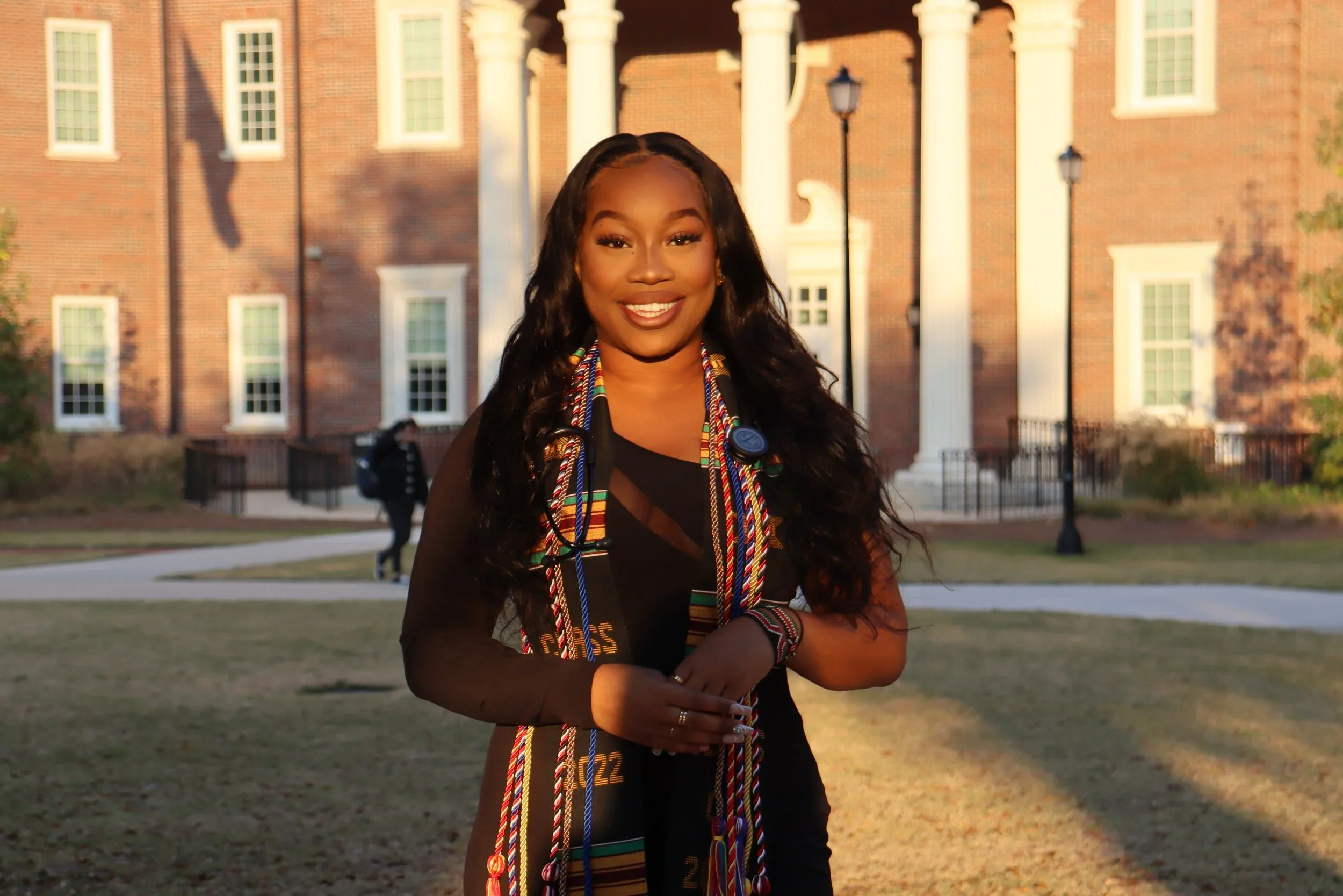 A smiling young woman in black outfit with a stethoscope around her neck and colorful graduation cords stands outside a brick building with white columns.