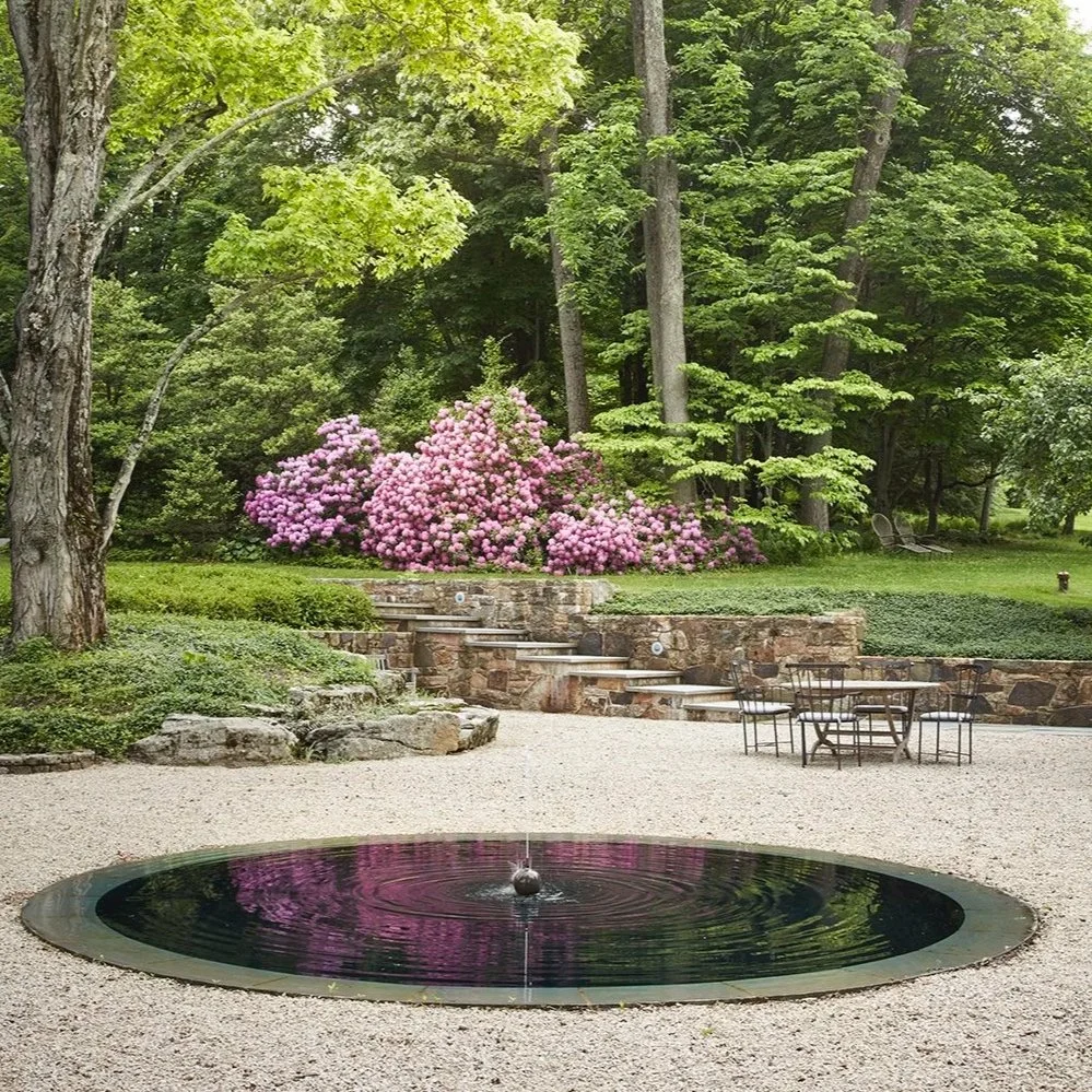 A peaceful garden scene with a round fountain with a small water feature, pink flowering bushes, green trees in the background, a stone wall with steps, and outdoor seating. Garden design by Fernando Caruncho and curated, gardened by Janet Mavec. 