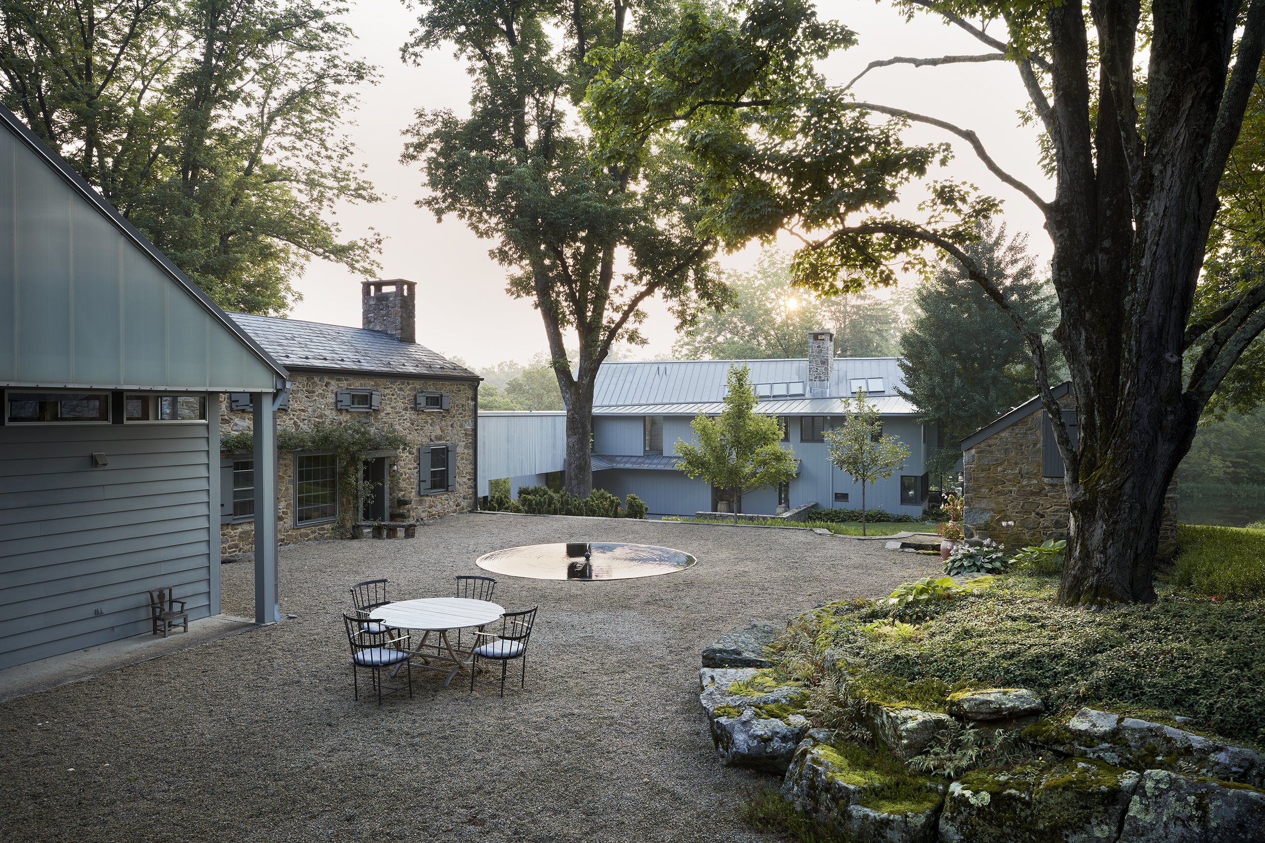 Sunshine filters through trees onto a cluster of residential buildings with stone and siding exteriors, a gravel courtyard, and outdoor furniture.