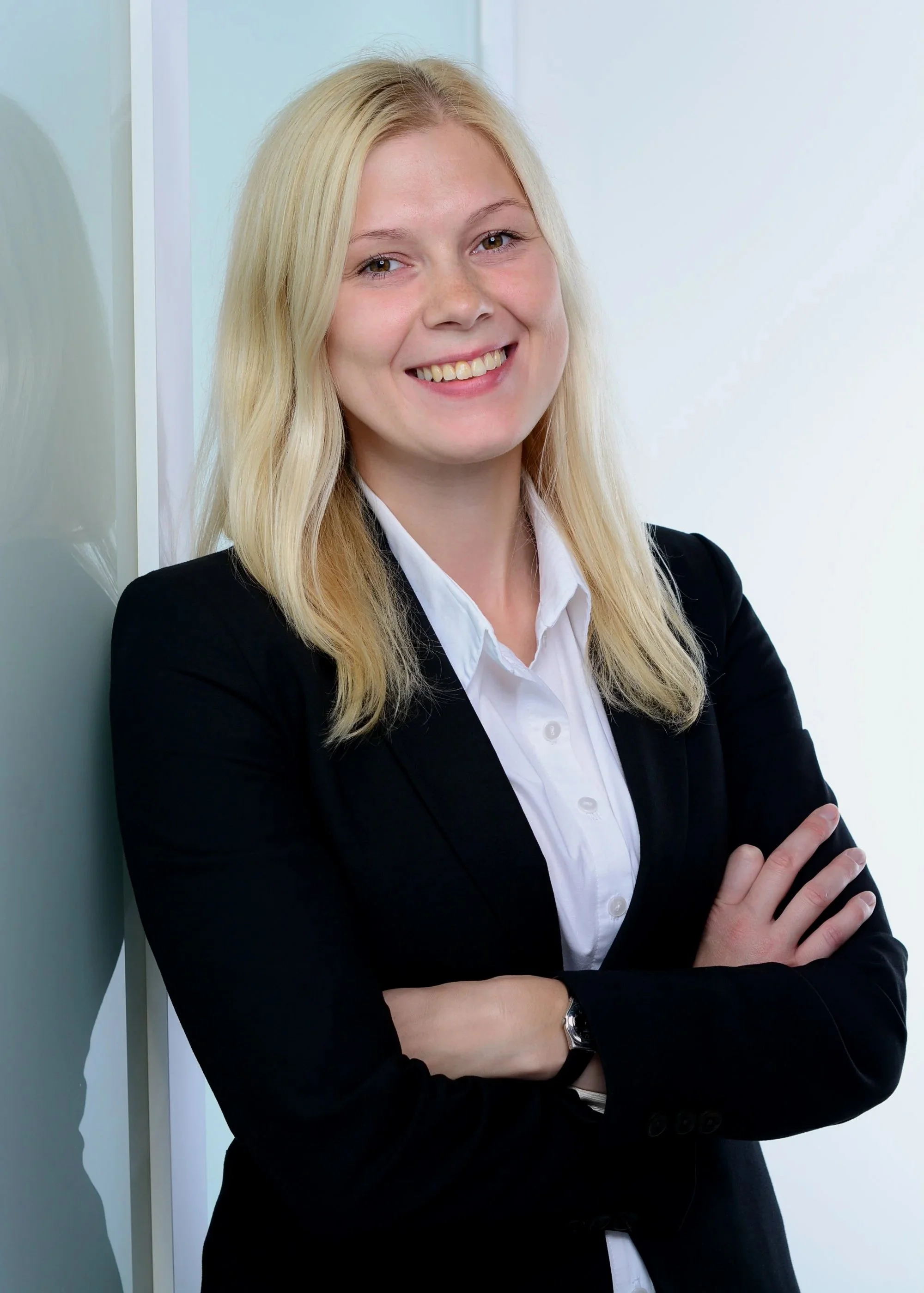 A young woman with long blond hair, wearing a black blazer and white shirt, is smiling and crossing her arms while standing against a light-colored wall.