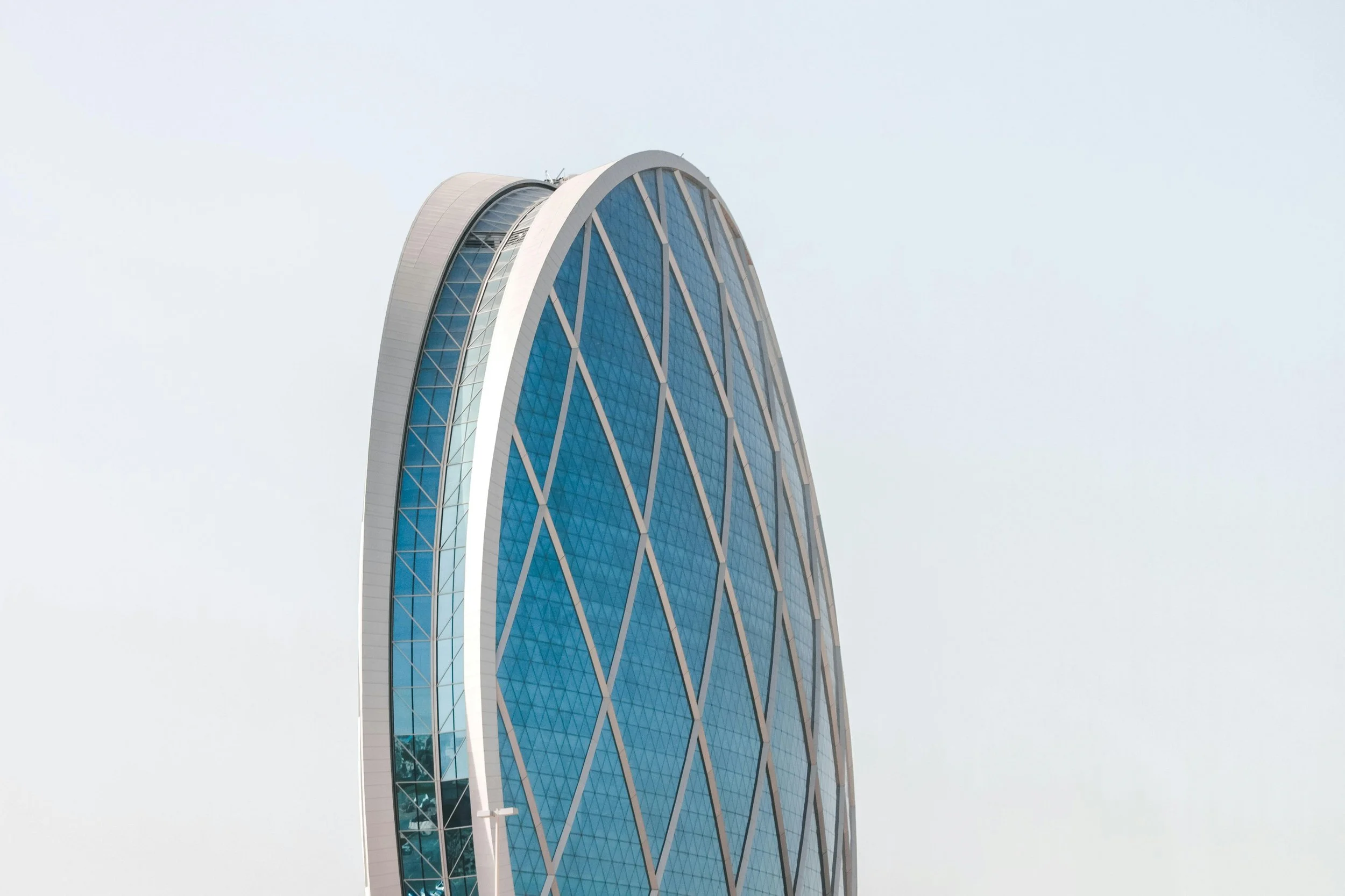 A modern skyscraper with a curved, sail-like shape covered in blue glass windows and white structural beams, set against a pale sky.