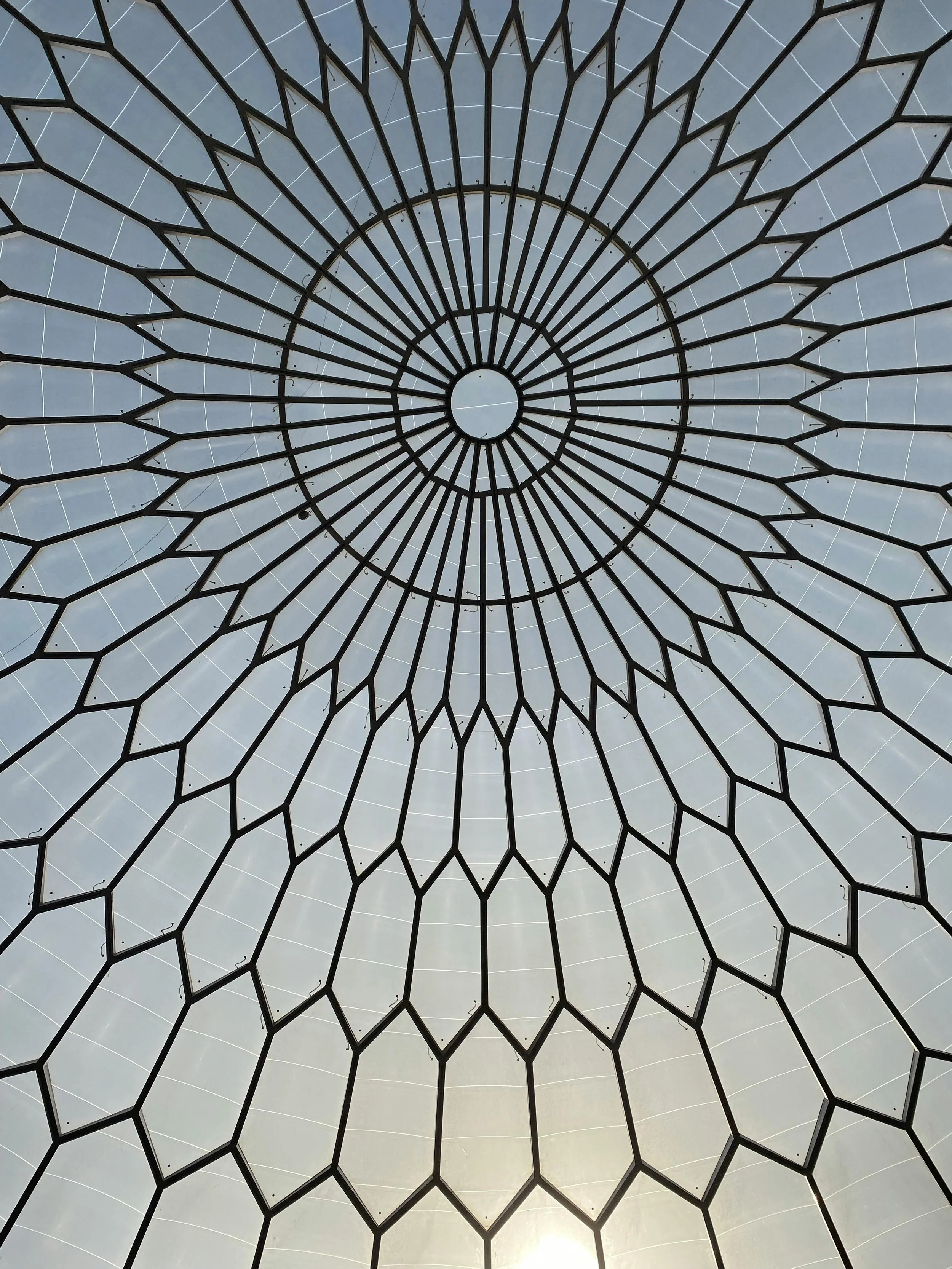 Looking up at a large, circular, geometric metal and glass dome structure with a central circular opening and radiating spokes.