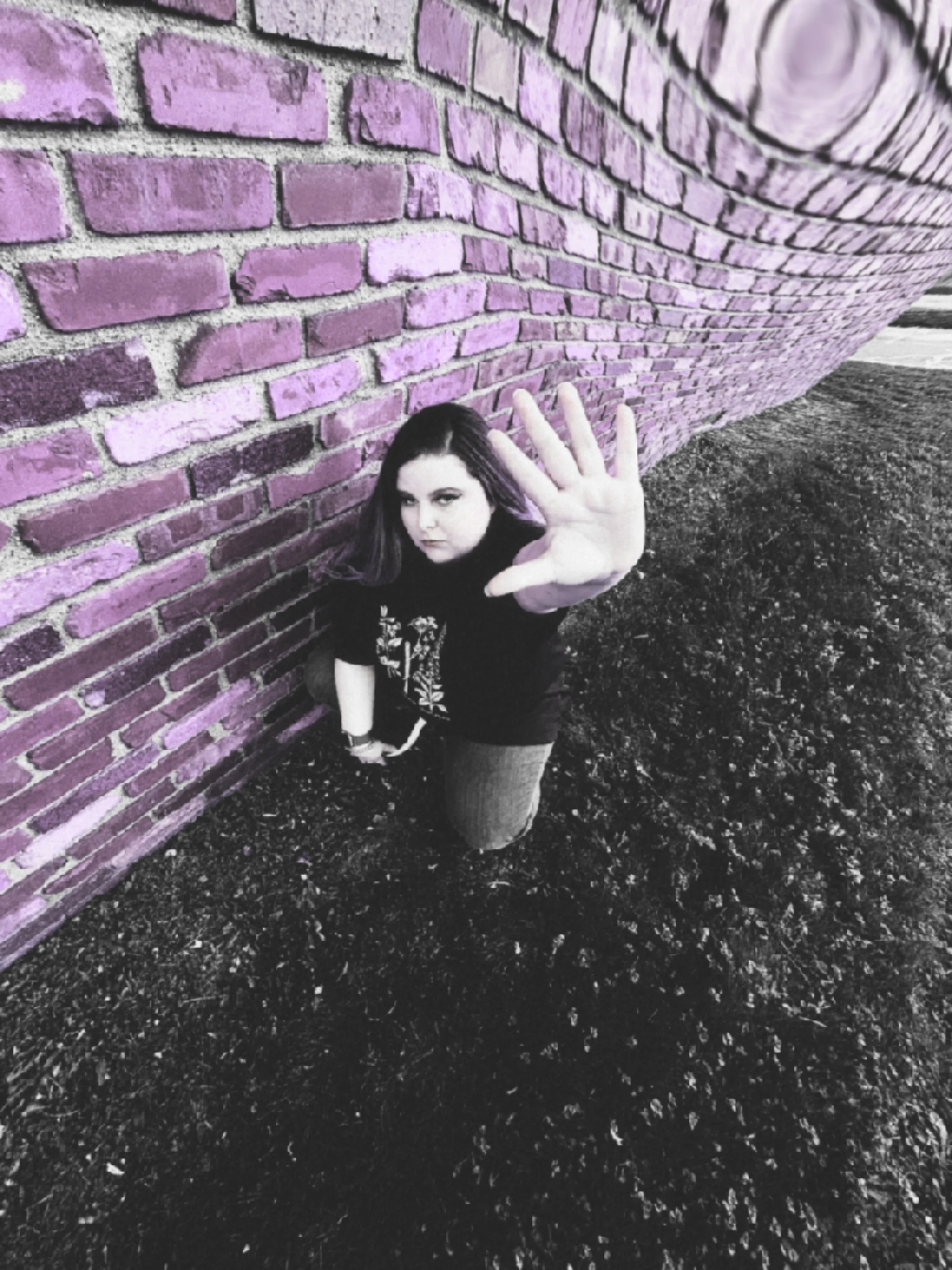 A woman standing on dark gravel ground next to a purple brick wall, reaching out with her hand towards the camera with an intense expression.