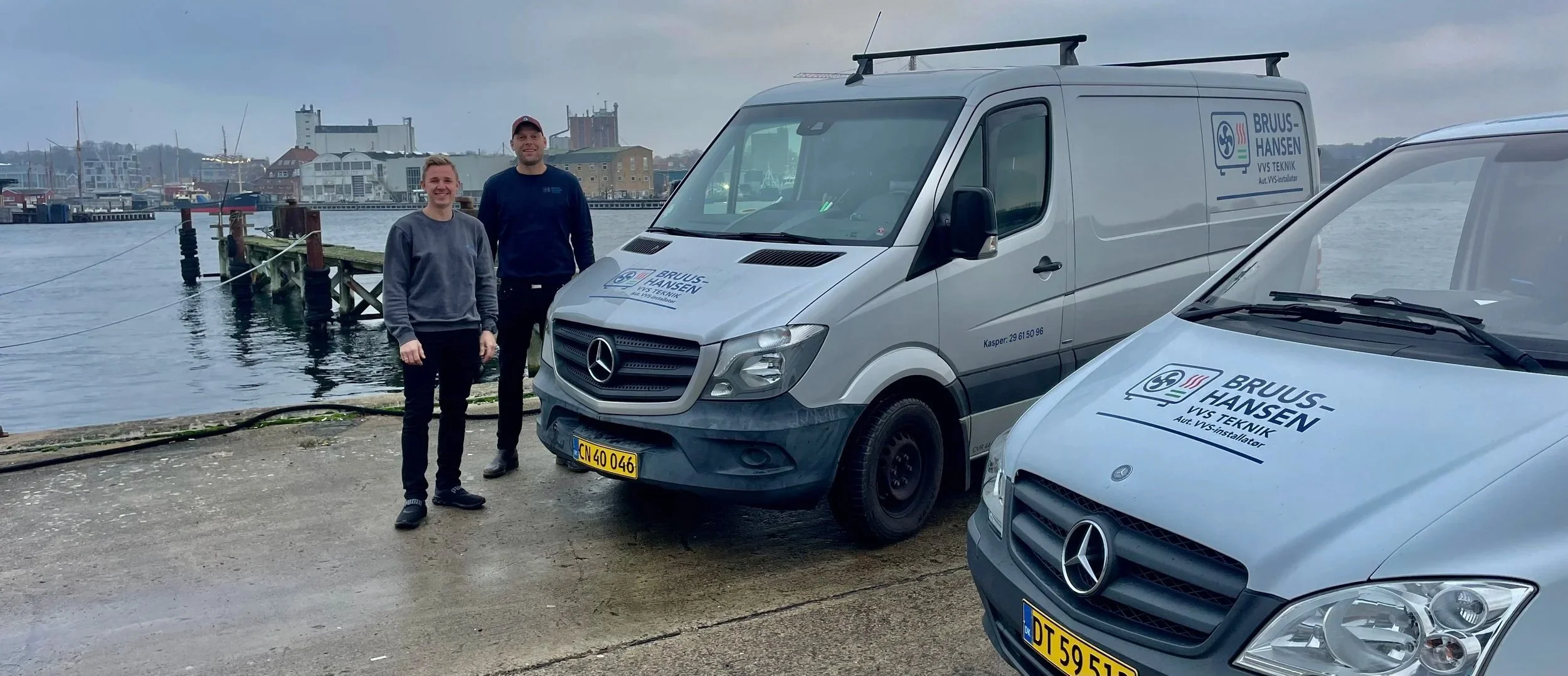 Two men standing next to two silver service vans with company logos parked near a body of water with boats and buildings in the background.