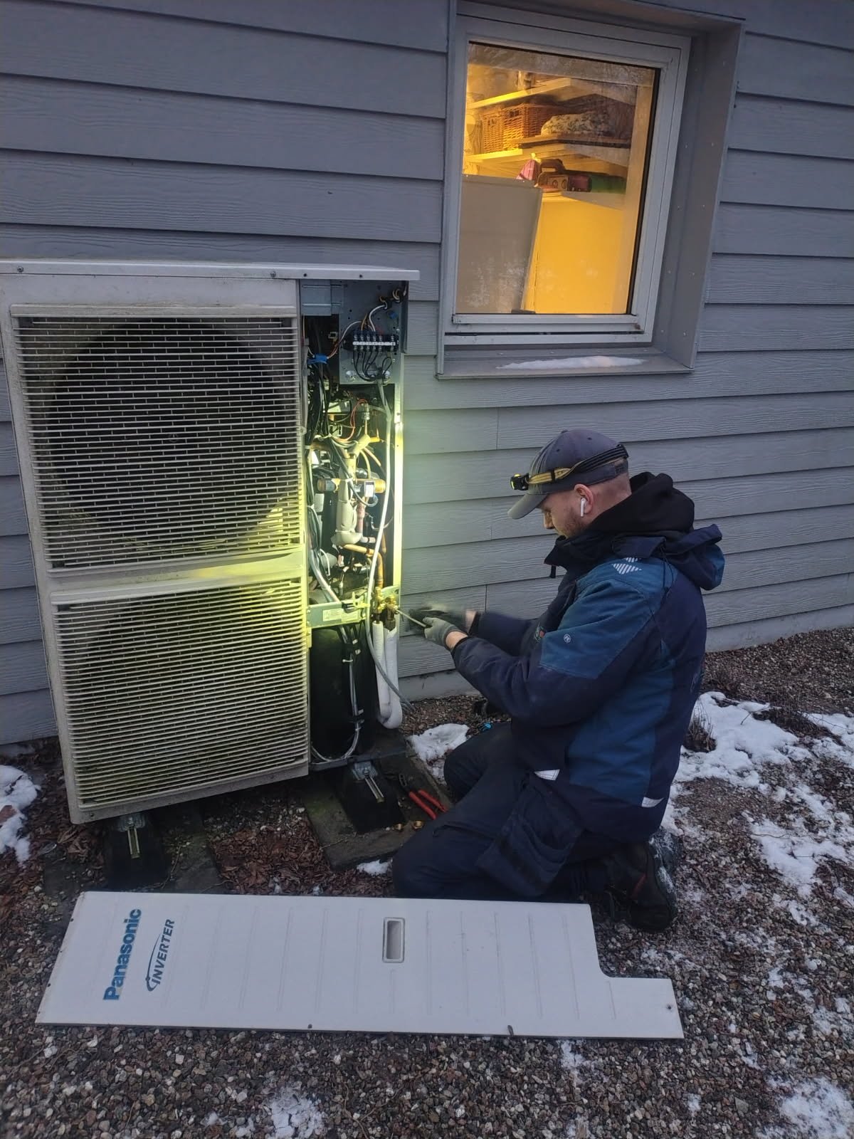 A technician repairing an outdoor air conditioning unit next to a house with gray siding, kneeling on the ground with tools, with a white Panasonic inverter panel on the ground and a window above showing the interior of the house.
