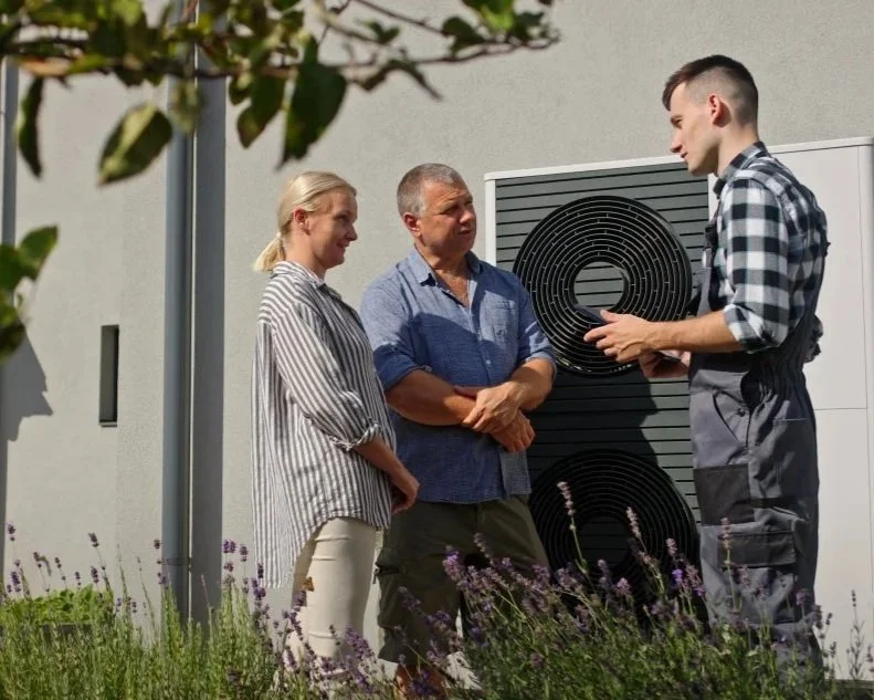 A man is explaining an HVAC unit to a woman and a man outdoors next to a building with purple flowers in the foreground.