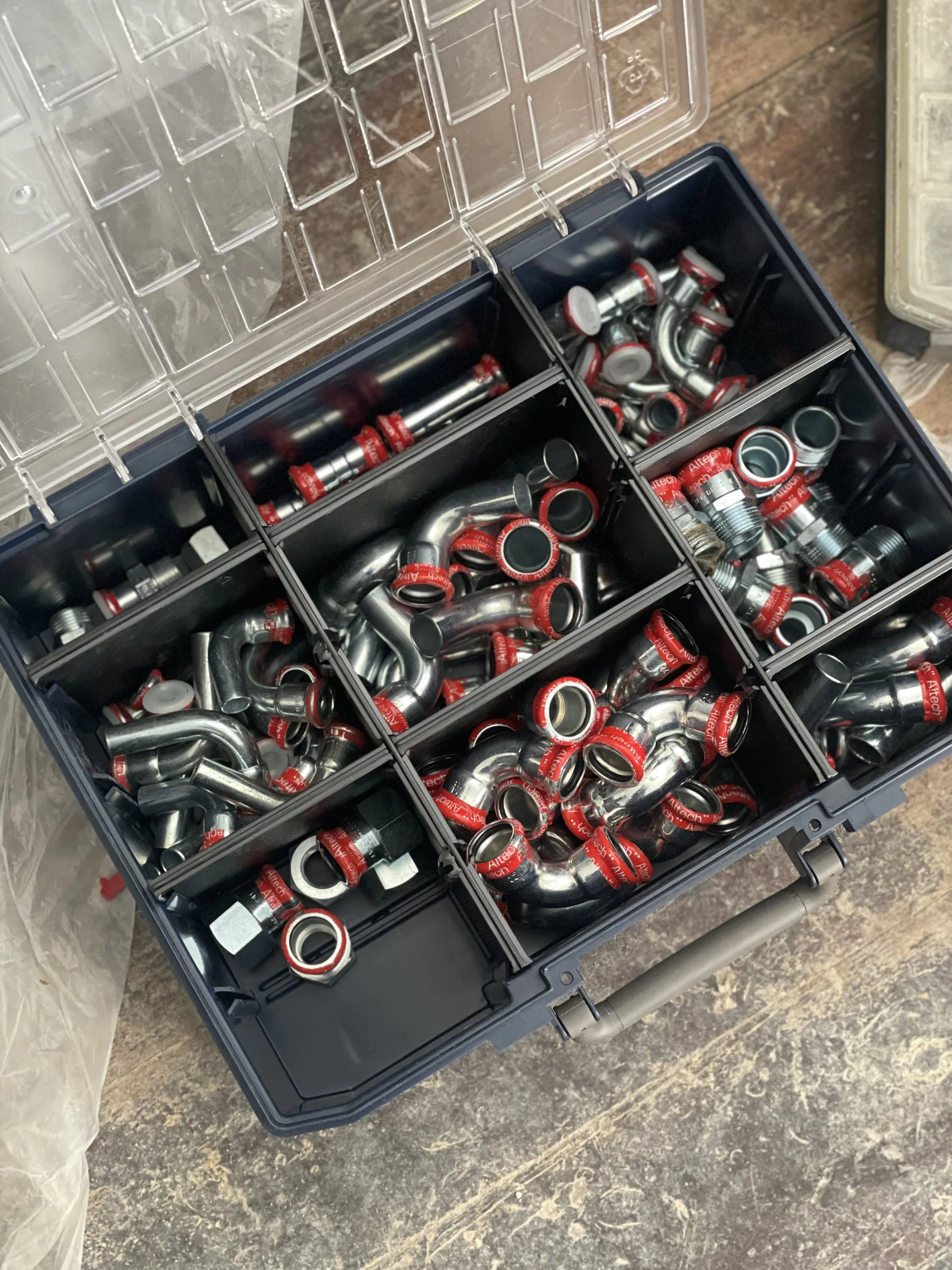 A black plastic storage box with multiple compartments filled with metal plumbing fittings, including elbows, couplings, and 90-degree bends, some with red labels and some plain.