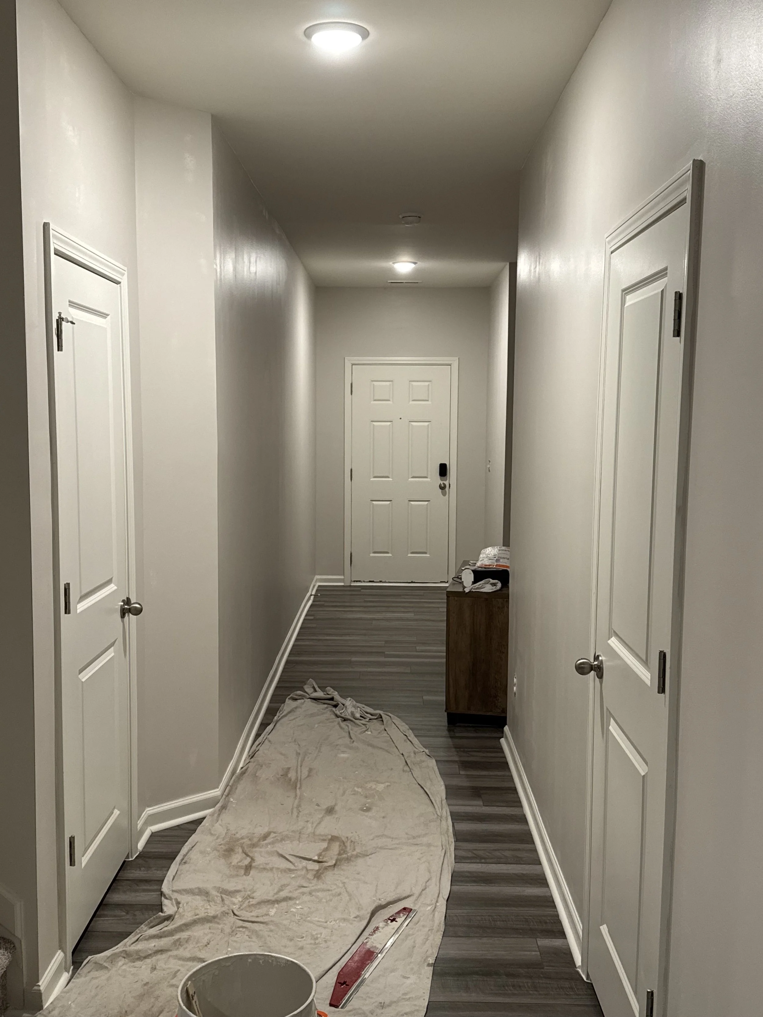 Empty hallway with three ceiling lights, two white closet doors on either side, a small wooden cabinet, and a white front door at the end. Construction or painting supplies on the floor, including a drop cloth, a paint bucket, and a level.