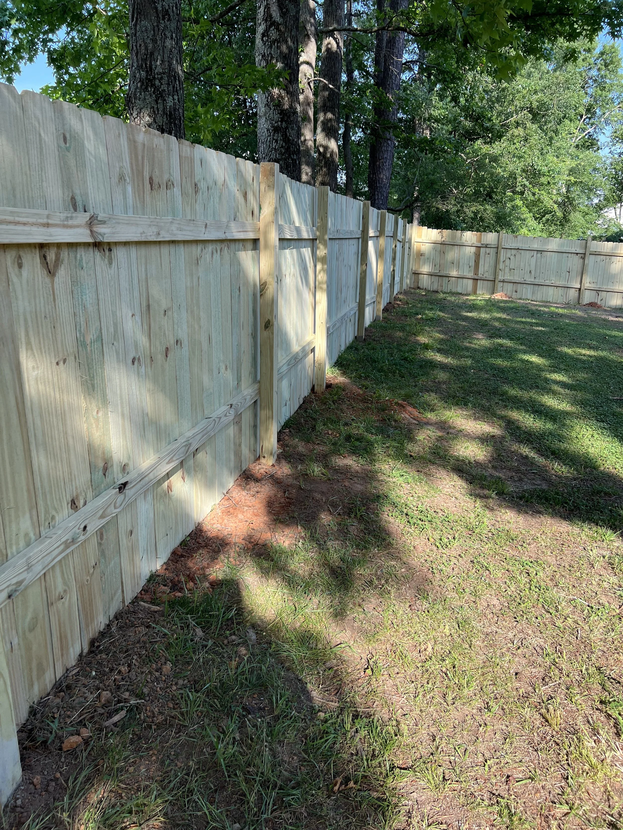 Wooden privacy fence in a backyard with grass and trees in the background, sunlight casting shadows on the ground.