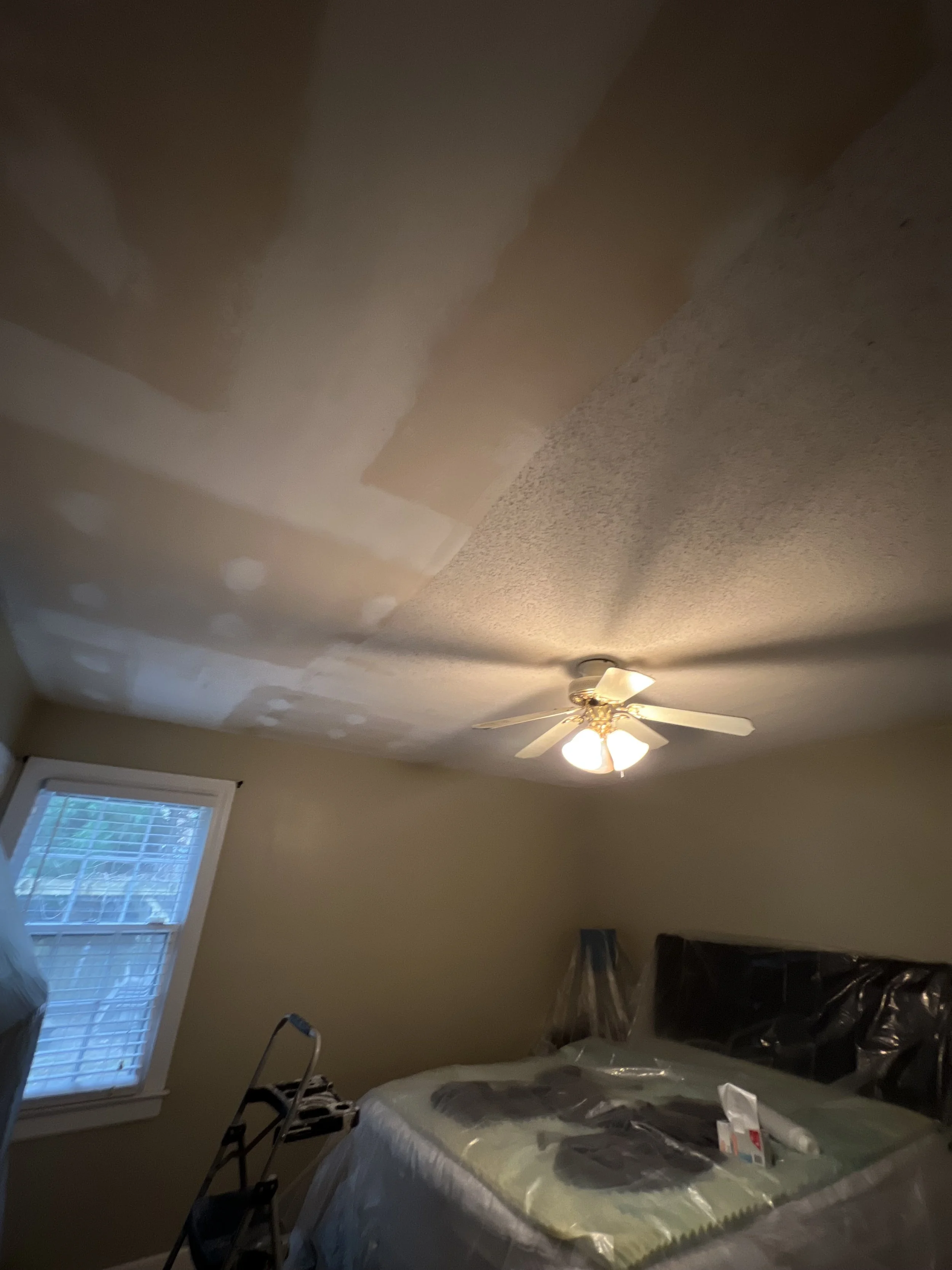 A bedroom ceiling with unfinished drywall and patches, a ceiling fan with lights, a window with blinds, a bed covered with a plastic sheet, and a step ladder in the corner.