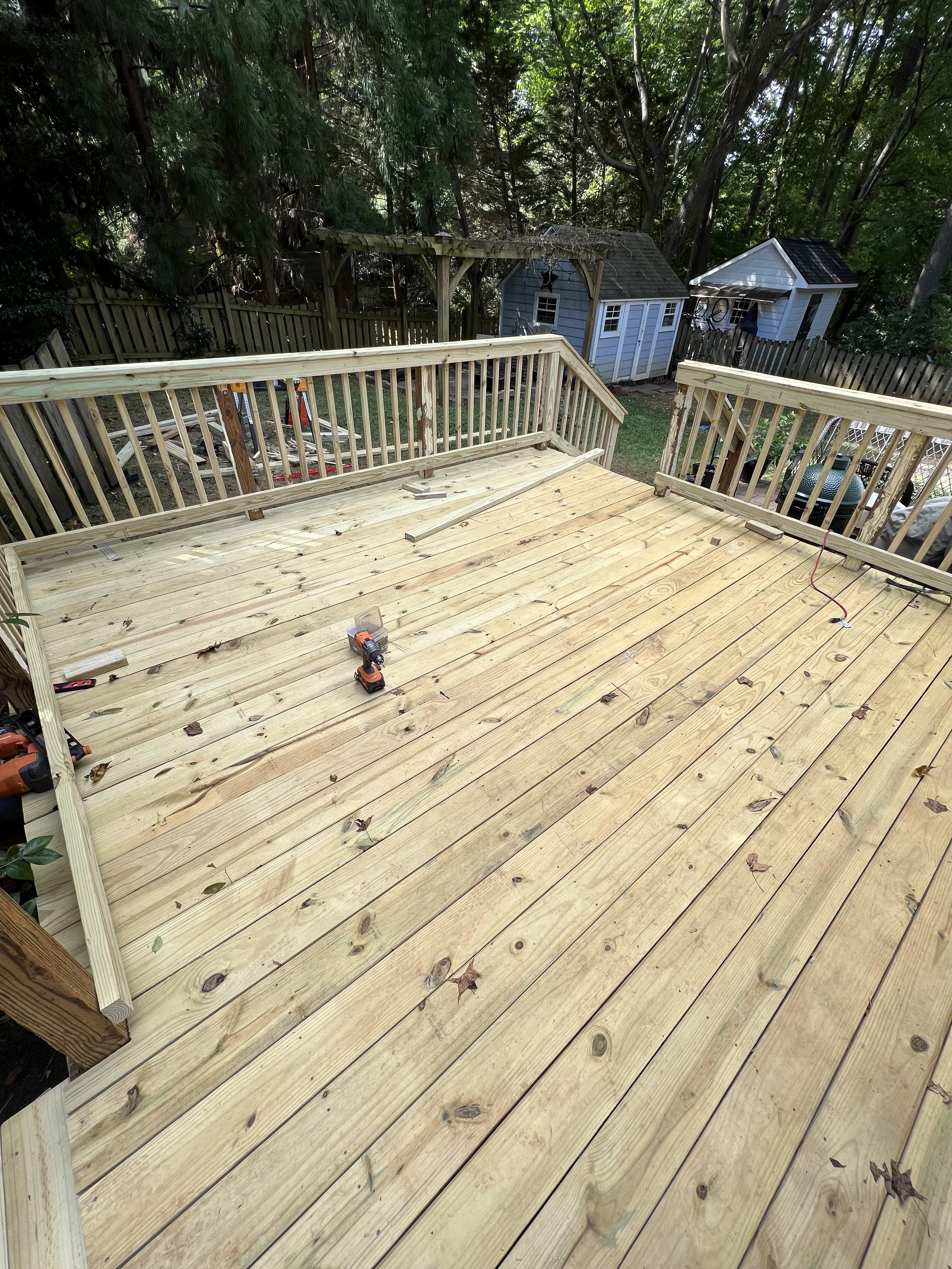 A wooden deck under construction in a backyard, with tools and leaves scattered on the surface, surrounded by trees and small sheds.
