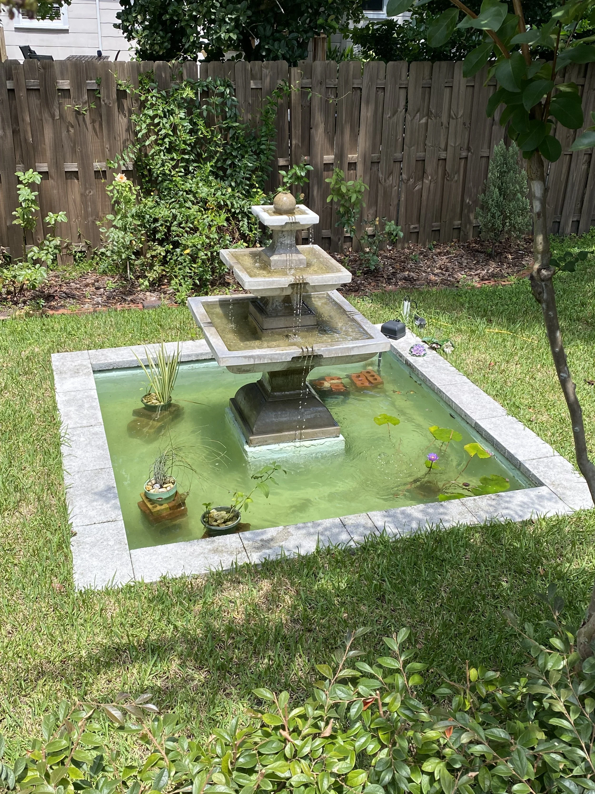 A backyard with a stone fountain in the center, surrounded by a grassy lawn and a wooden fence. The fountain has three tiers with water streaming from the top and features small potted plants around its base. There are various bushes and trees along 
