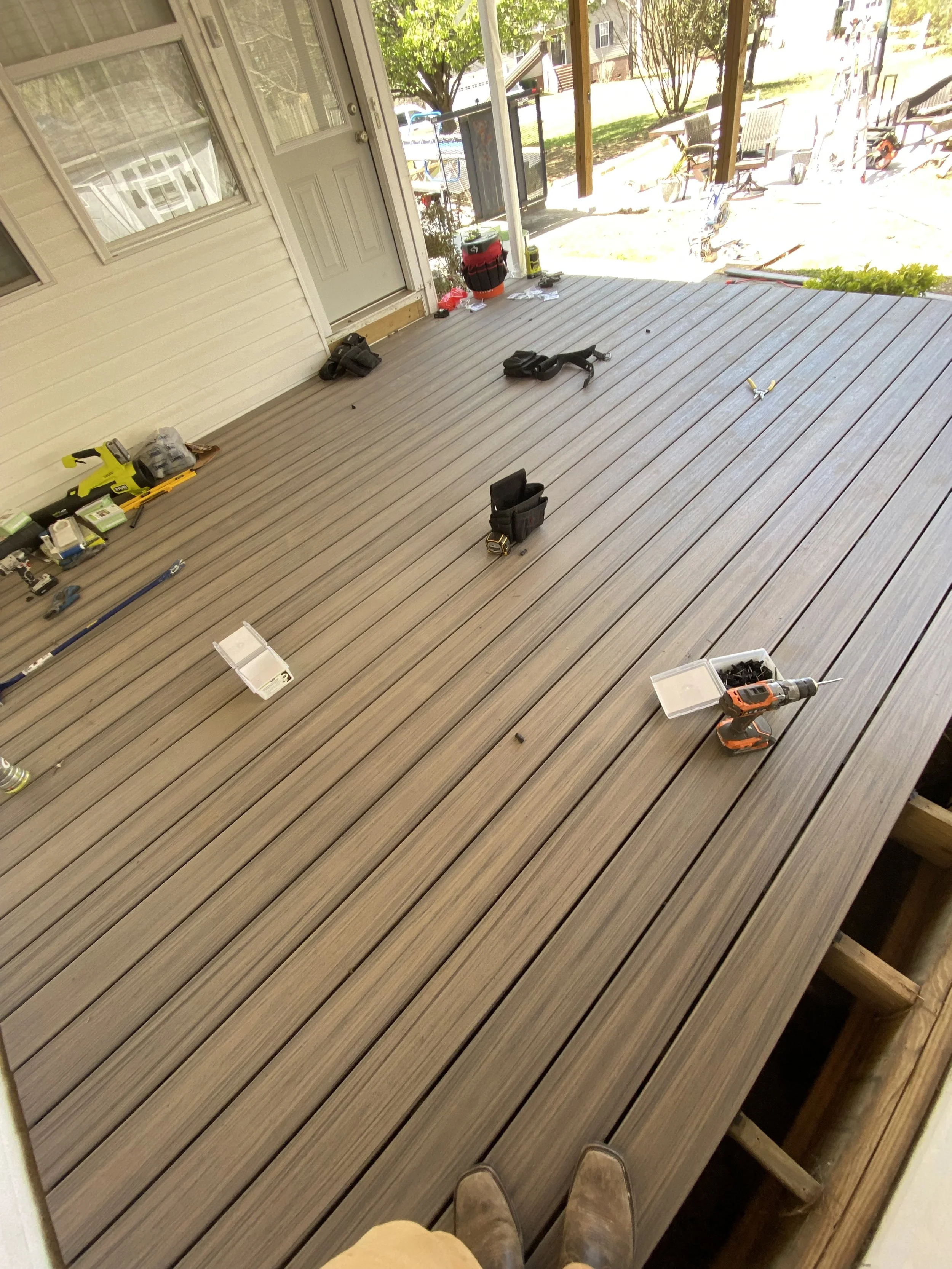 View of a porch or deck with construction tools and supplies scattered around, including a cordless drill, tool pouches, and boxes, with a view outside showing trees and a yard.