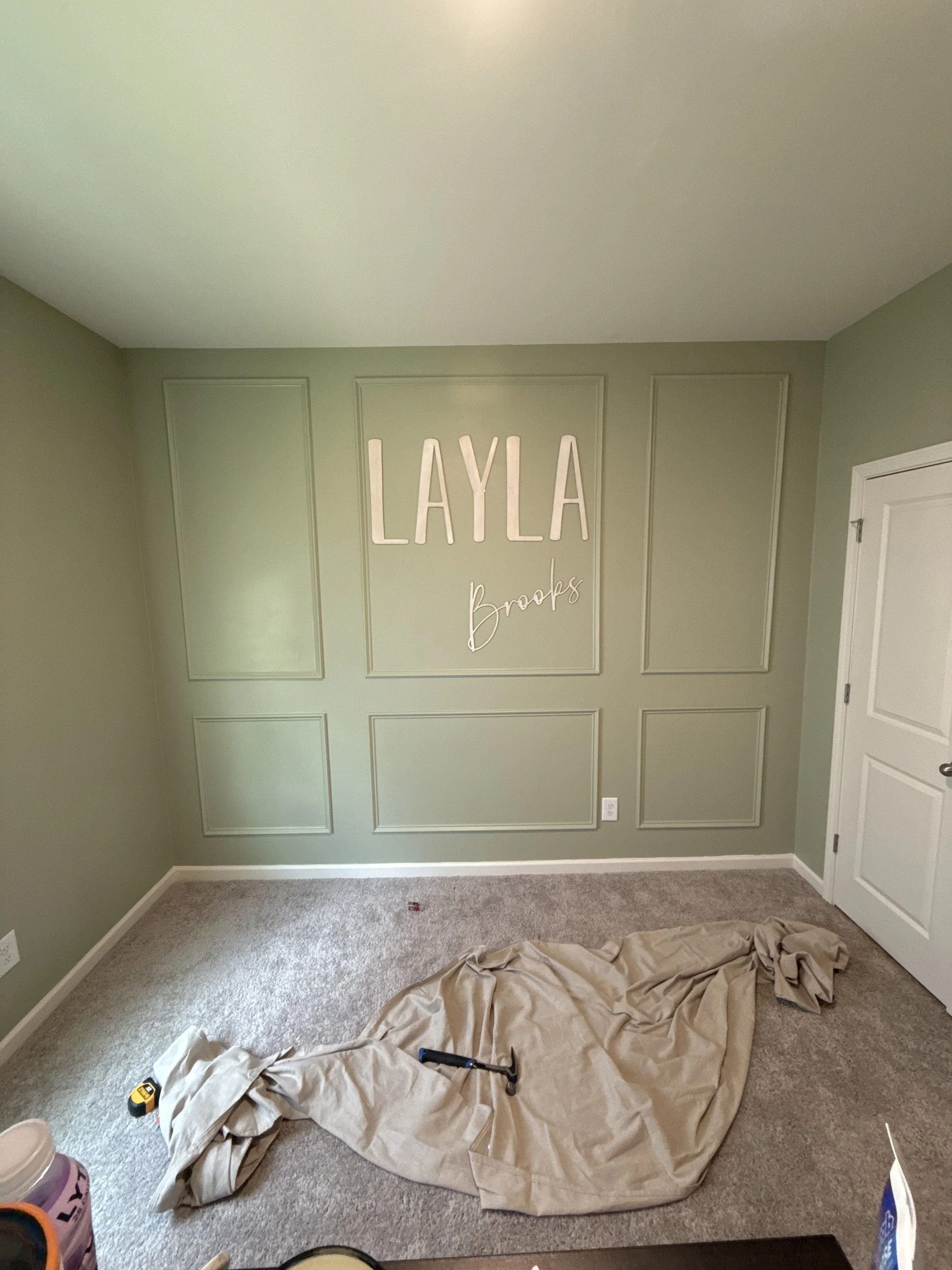 Room with a green wall, white decorative wall panels, and oversized white letters spelling 'LAYLA' above a smaller cursive 'Brooks.' There is a beige cloth on the carpeted floor, along with a hammer, measuring tape, and a purple bucket.