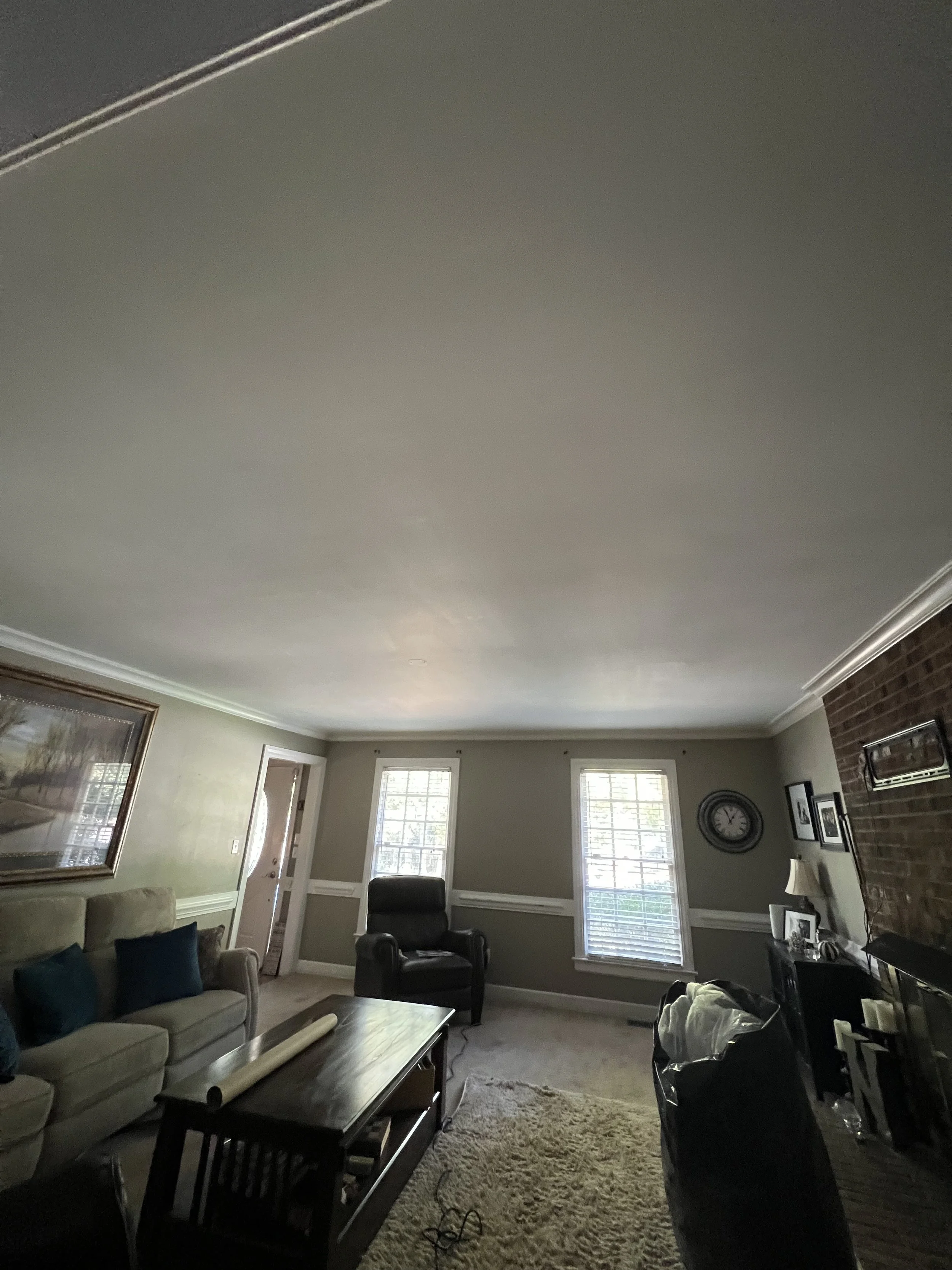 Living room with beige sofa, black armchair, coffee table, window blinds, brick fireplace, wall clock, framed pictures, and a lamp.