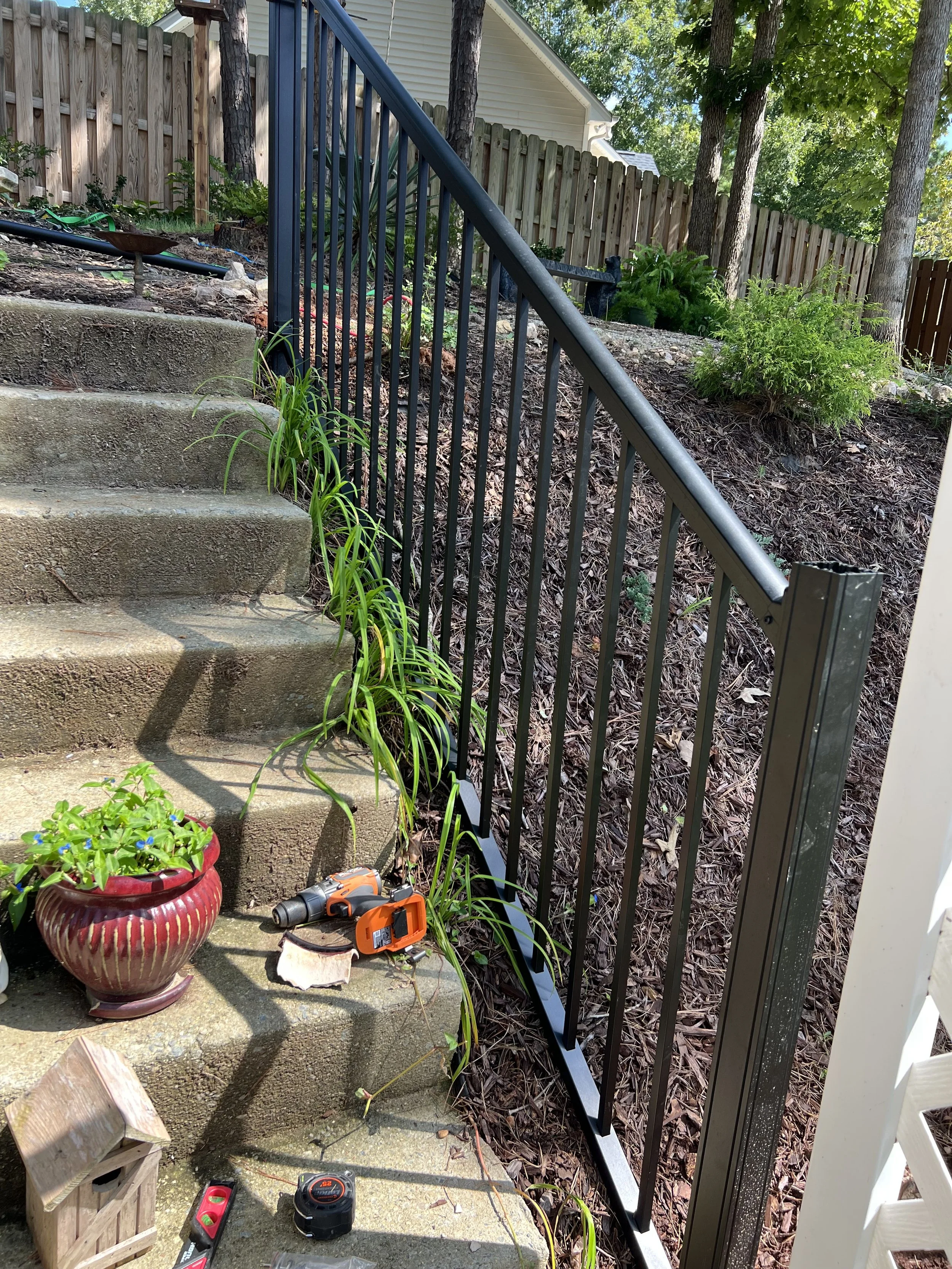 Outdoor scene showing concrete steps, a black metal handrail, potted plants, a toolbox, electric drill, measuring tape, and other tools. A backyard with trees and a wooden fence in the background.
