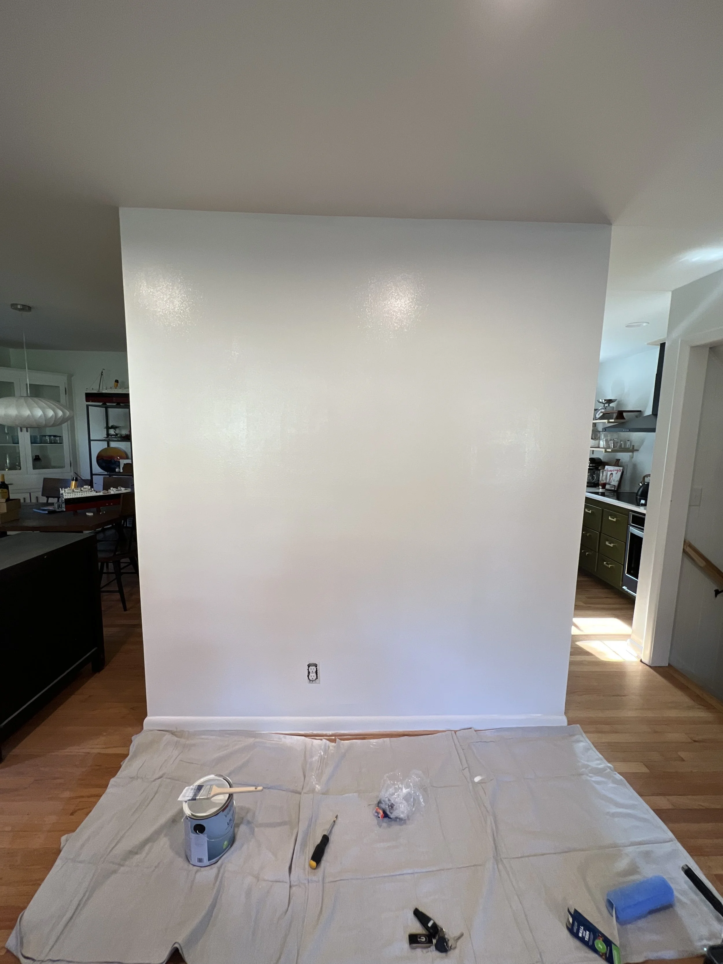 A partially painted white wall in a room with painting tools and supplies on the floor, including a paint can, brush, roller, and tape, with a protective cloth covering the floor.