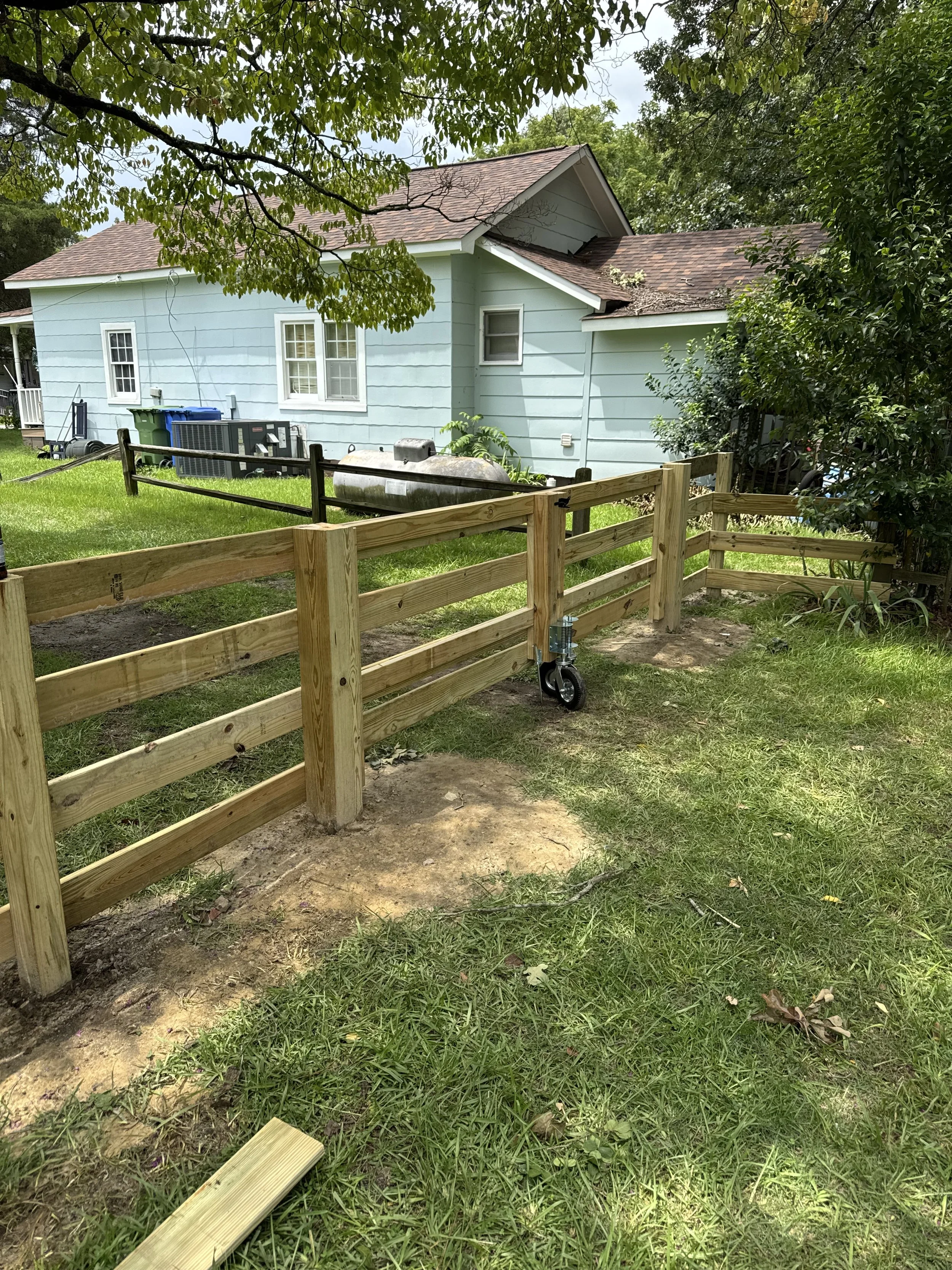 A partially built wooden fence inside a backyard, with a house and green trees in the background.