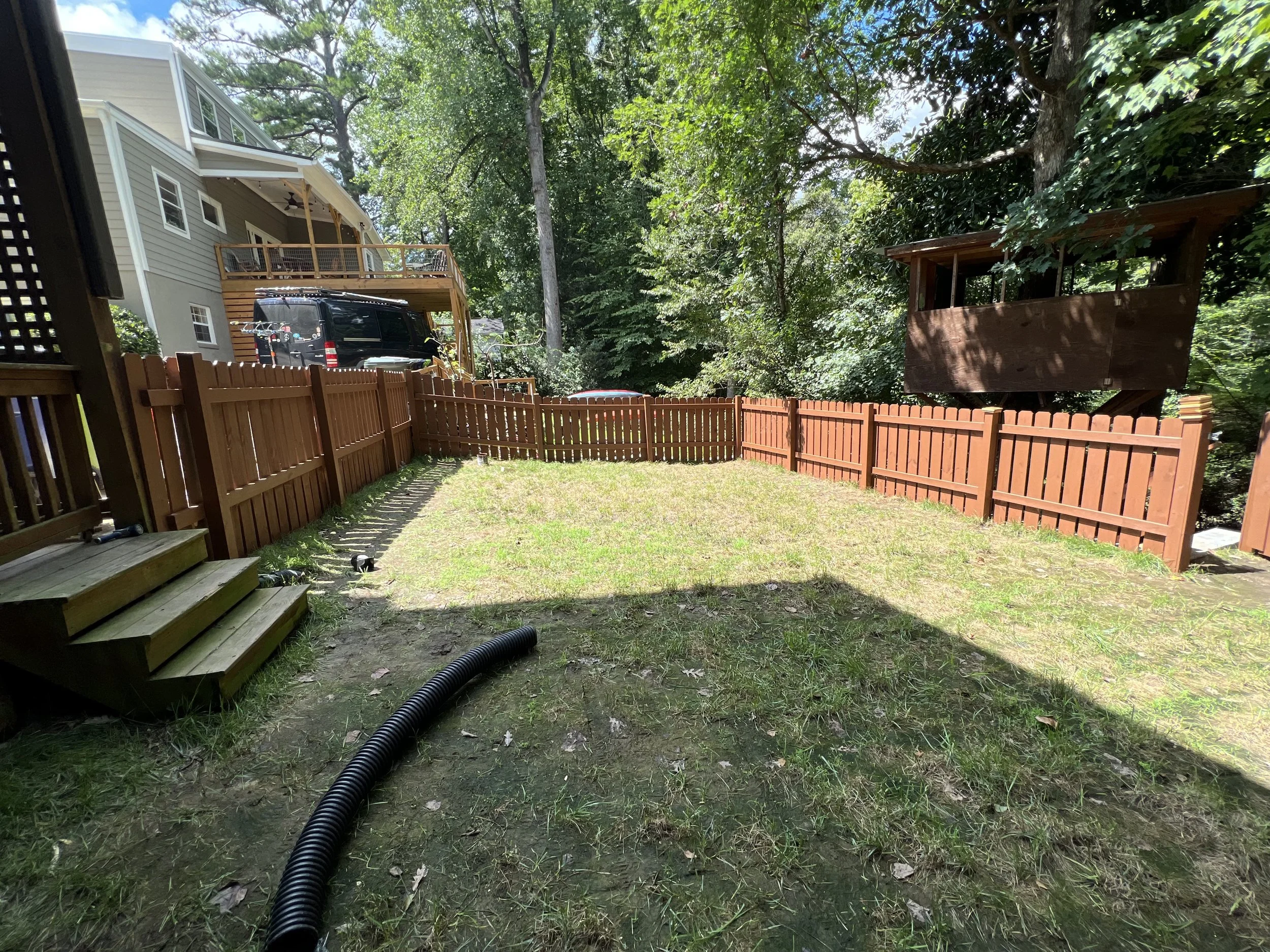 Backyard with a green lawn, a wooden fence, surrounded by trees, and a view of neighboring houses and a treehouse.