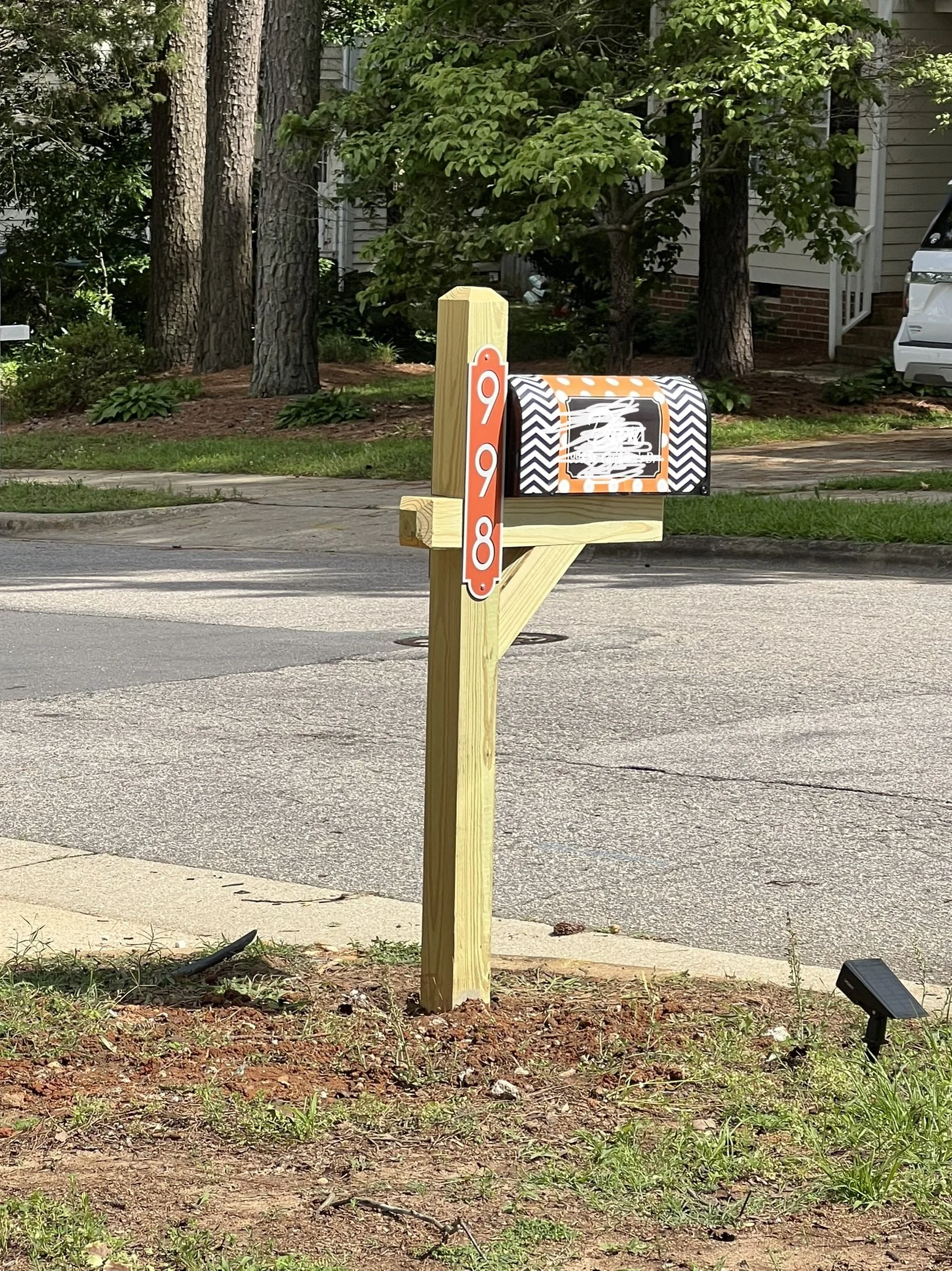 A mailbox mounted on a wooden post with a sign attached displaying the number 998. The mailbox has a black and white zigzag pattern with orange accents. The mailbox is on the edge of a sidewalk, with grass, trees, and residential houses visible in th
