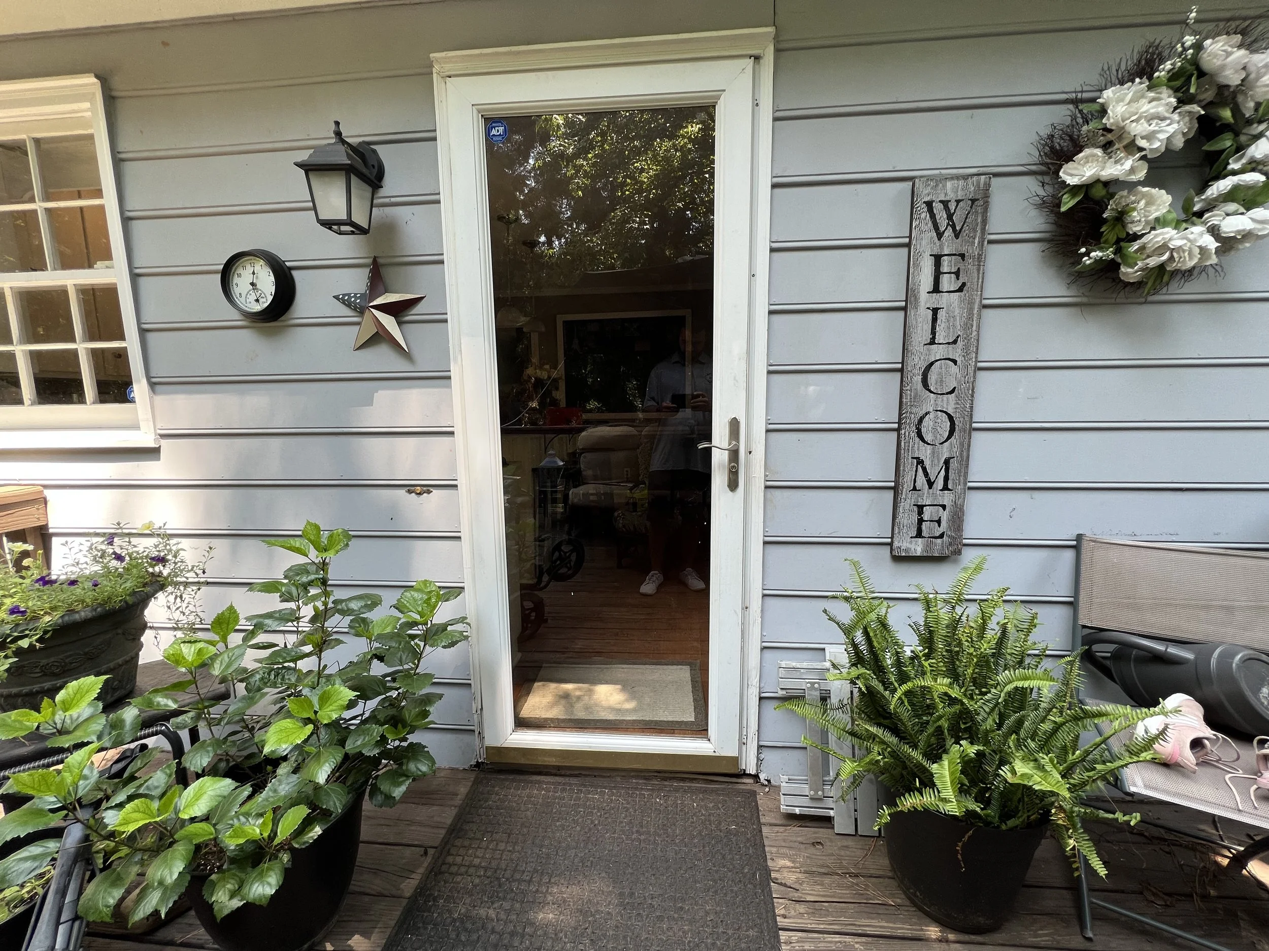 Front porch of a house with a glass door, a welcome sign, a flower wreath, potted plants, and outdoor decorations.