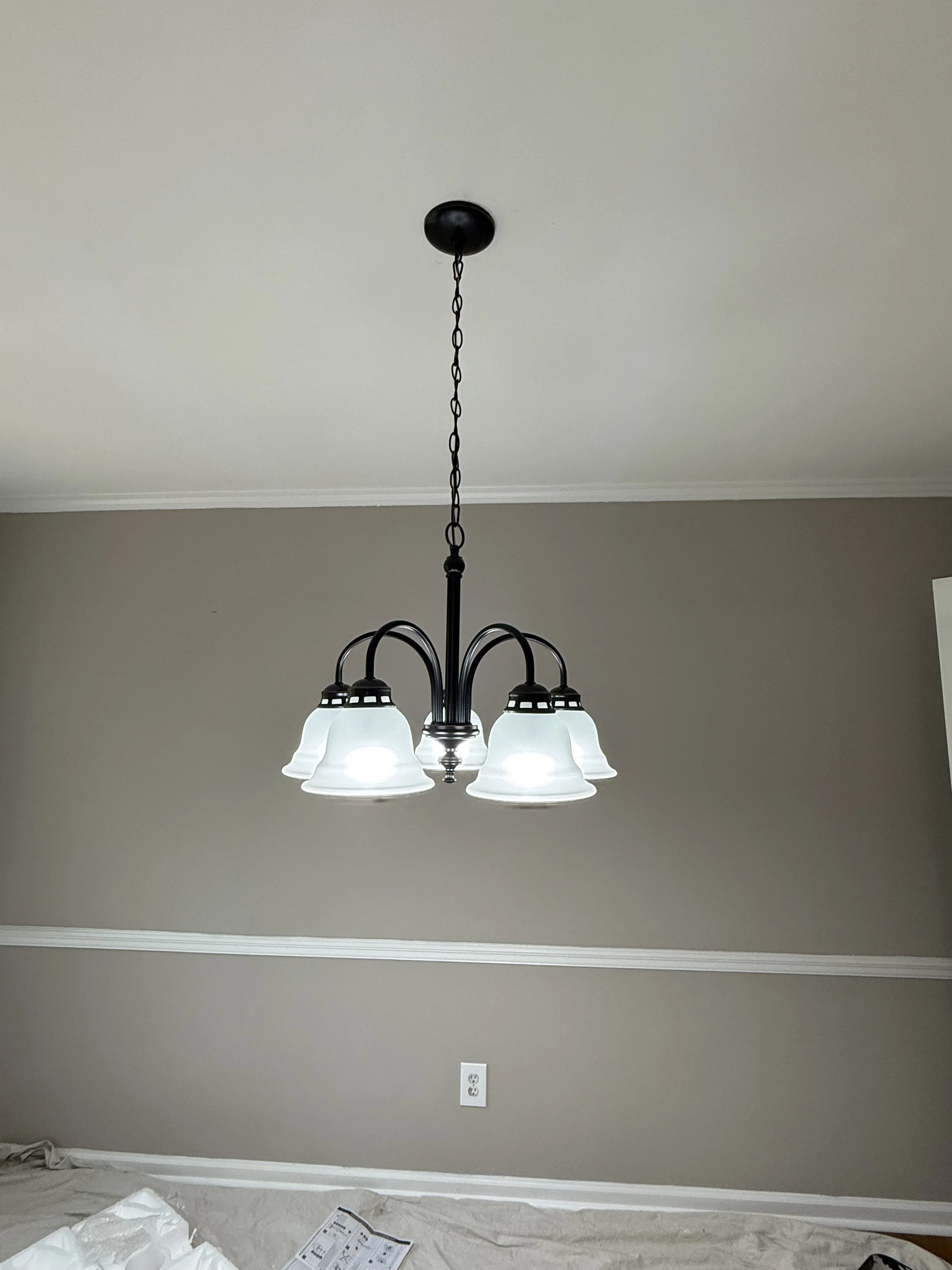 A black chandelier with five white glass shades hanging from a chain in a room with beige walls and a white electrical outlet.