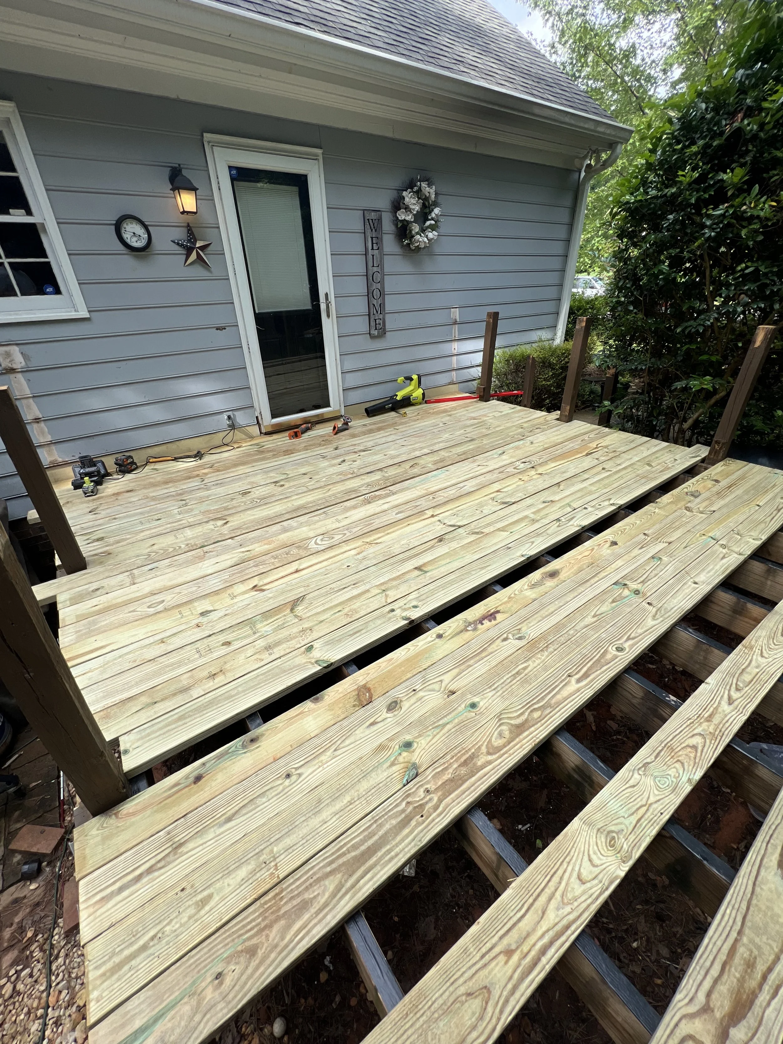 A backyard deck under construction with partially built wooden floorboards, tools and construction materials visible, attached to a house with gray siding, a glass door, and decorative outdoor wall hangings.