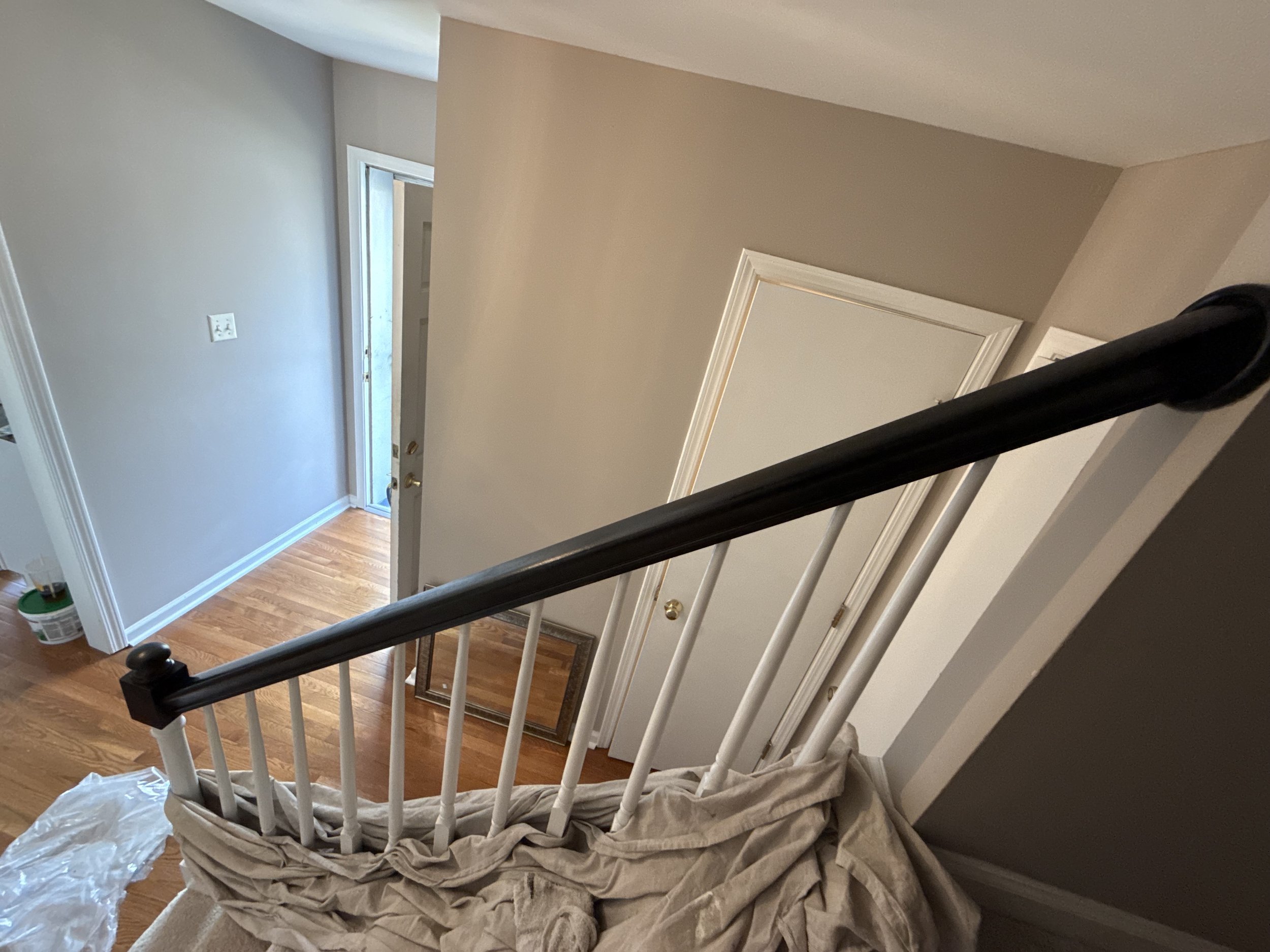 Inside view of a staircase with a black handrail and white spindles, with beige and gray walls, wood flooring, and a white door leading outside.