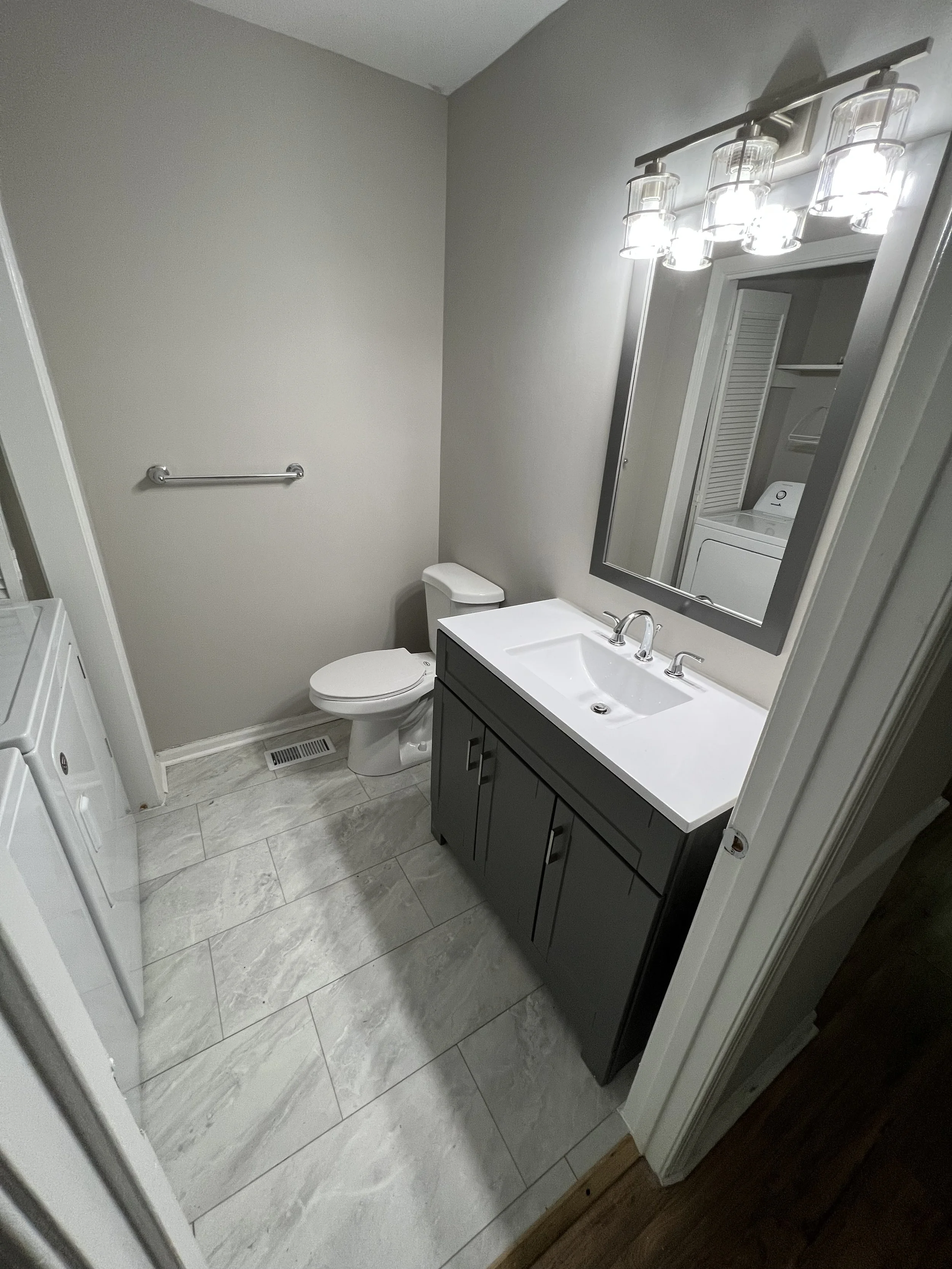A modern bathroom with a gray vanity, white sink, large mirror, chrome fixtures, light fixture above mirror, toilet, and a laundry area with a washing machine.