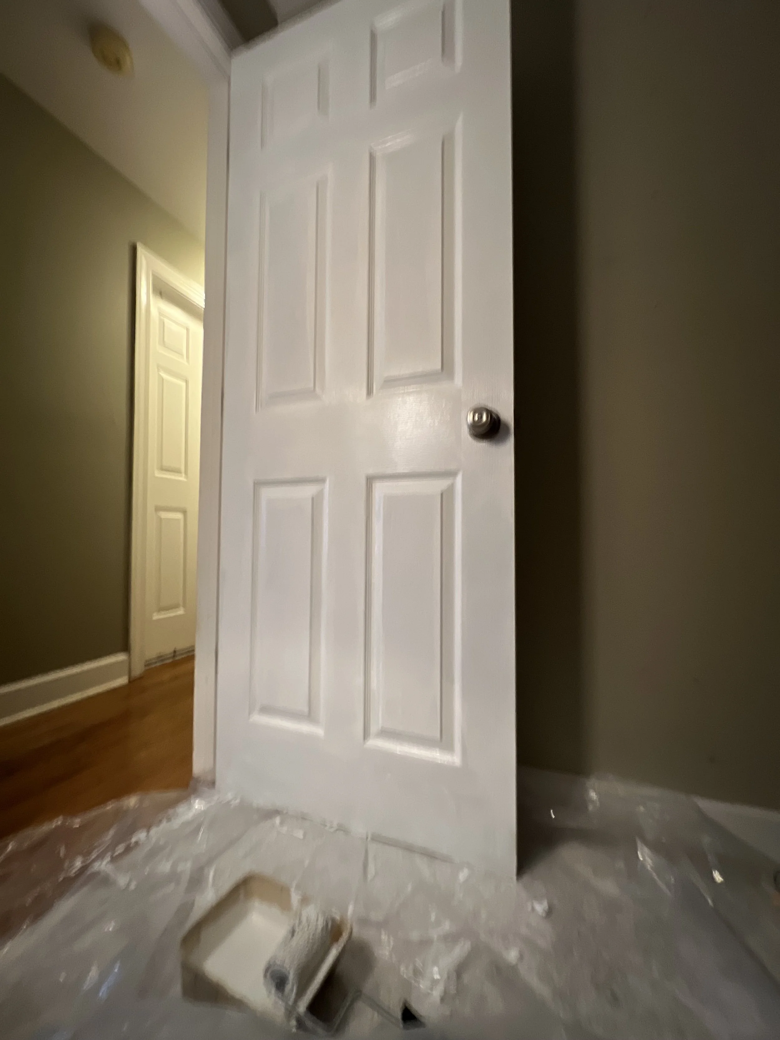 An interior hallway with a white six-panel door partially open, paint supplies on the floor, and plastic sheeting covering the floor.