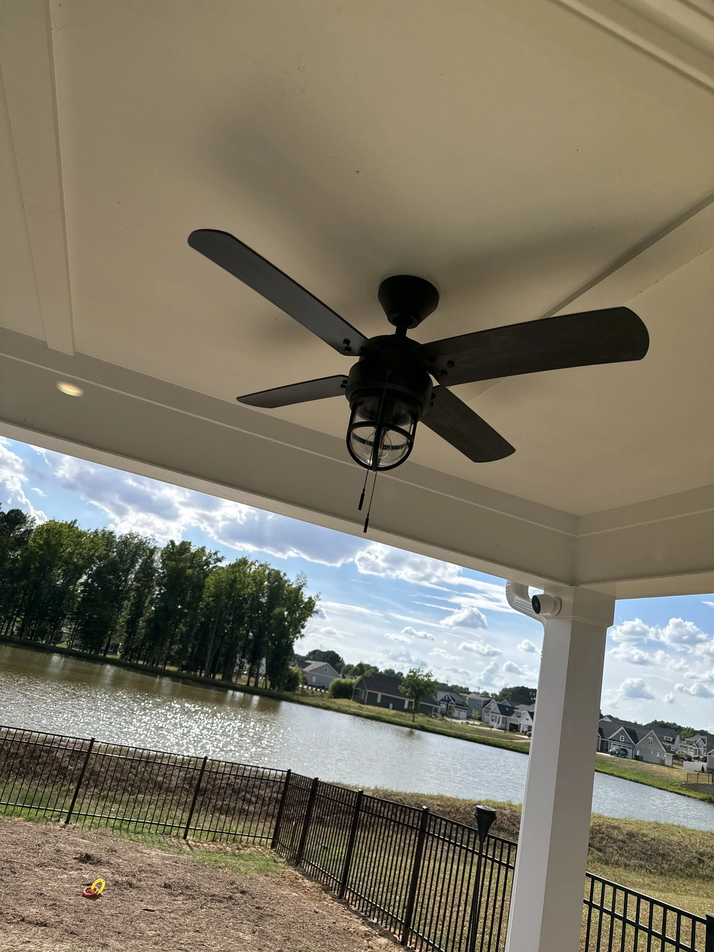 A ceiling fan with four black blades and a cage-style light fixture is mounted on a ceiling. Outside, there is a lake with trees on the far side, and a neighborhood with houses lining the street. The sky is partly cloudy with patches of blue visible.