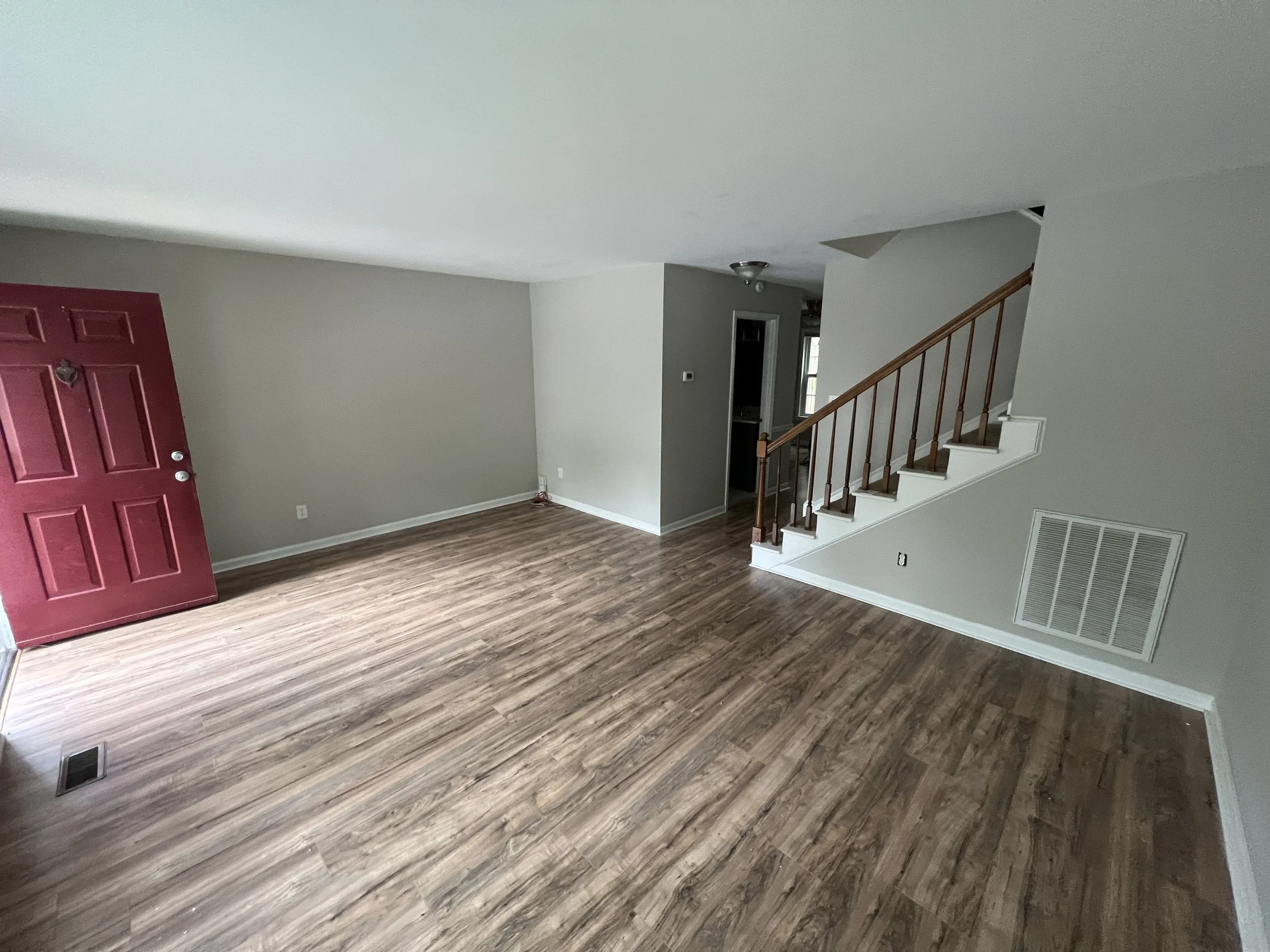 Empty living room with wooden floors, a red front door, staircase with wooden railing, gray walls, and a vent on the wall.
