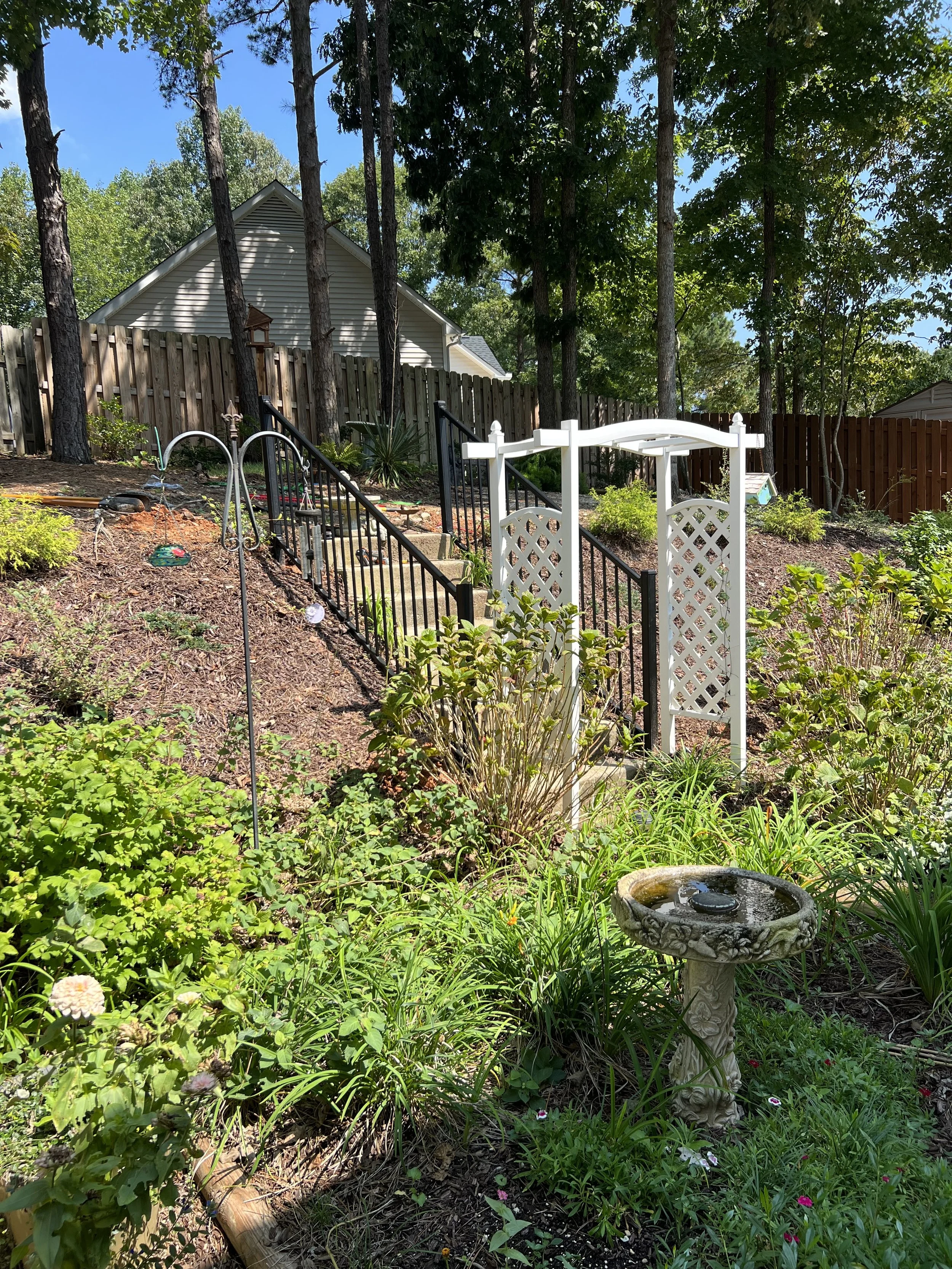 A backyard garden with a stone birdbath, lush green plants, a white decorative garden arch, and a black metal staircase leading up to a house behind a wooden fence, with tall trees and a blue sky in the background.
