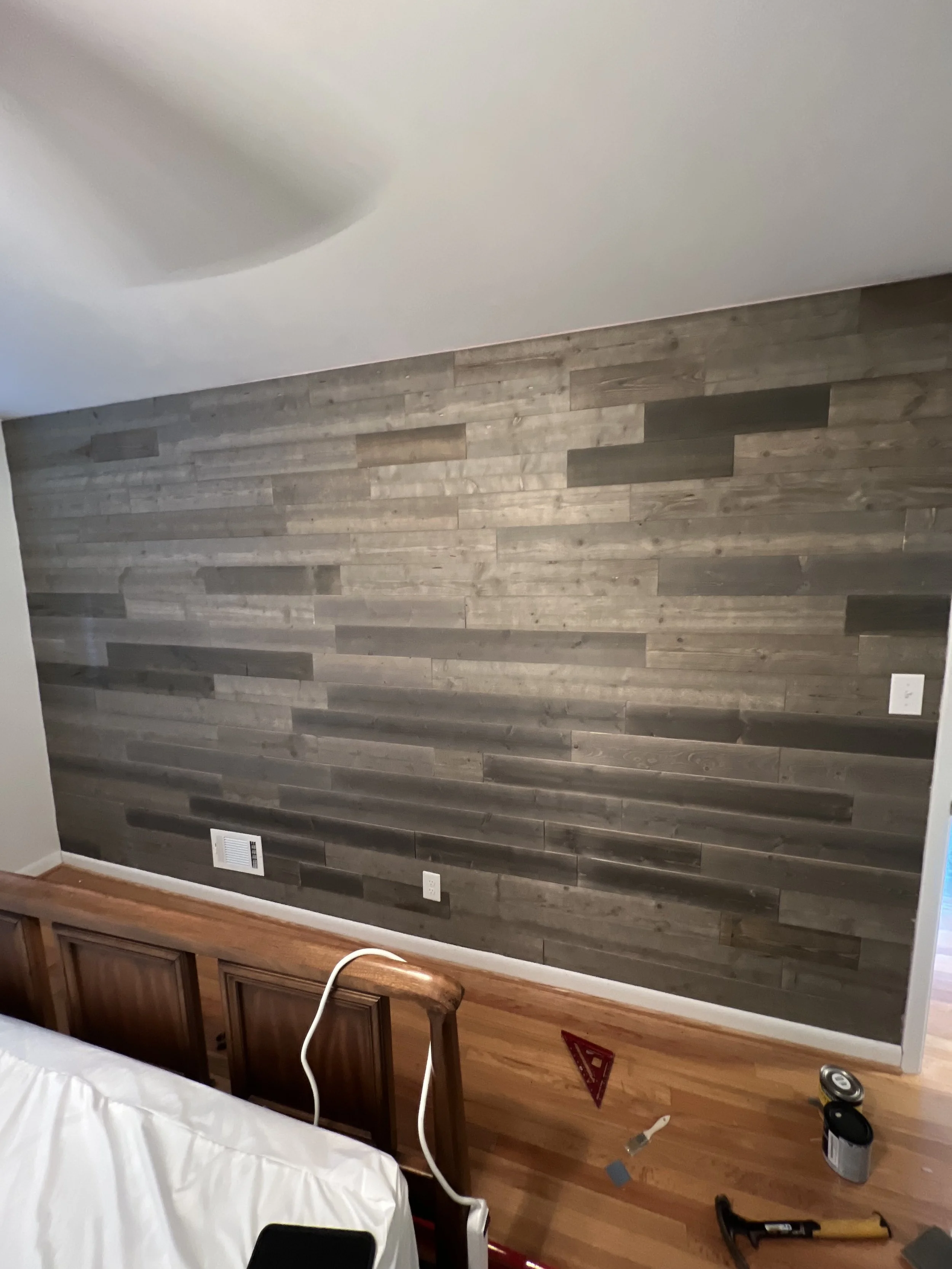 Interior of a room with a gray wooden accent wall, hardwood floor, and a white bed with a wooden headboard. Tools and a paint can are on the floor, indicating ongoing renovation.