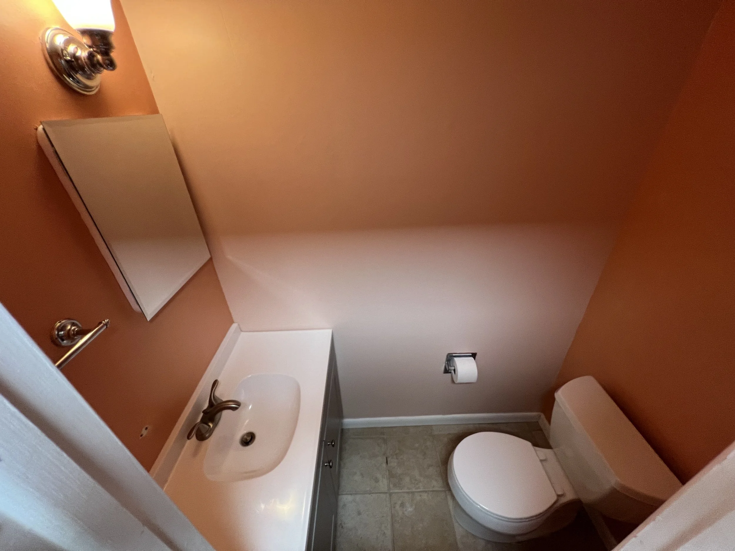 A small bathroom with peach-colored walls, a white toilet, a white sink with a cabinet below, a rectangular mirror above the sink, a wall-mounted towel ring, and a wall-mounted toilet paper holder. The floor is tiled with beige tiles.