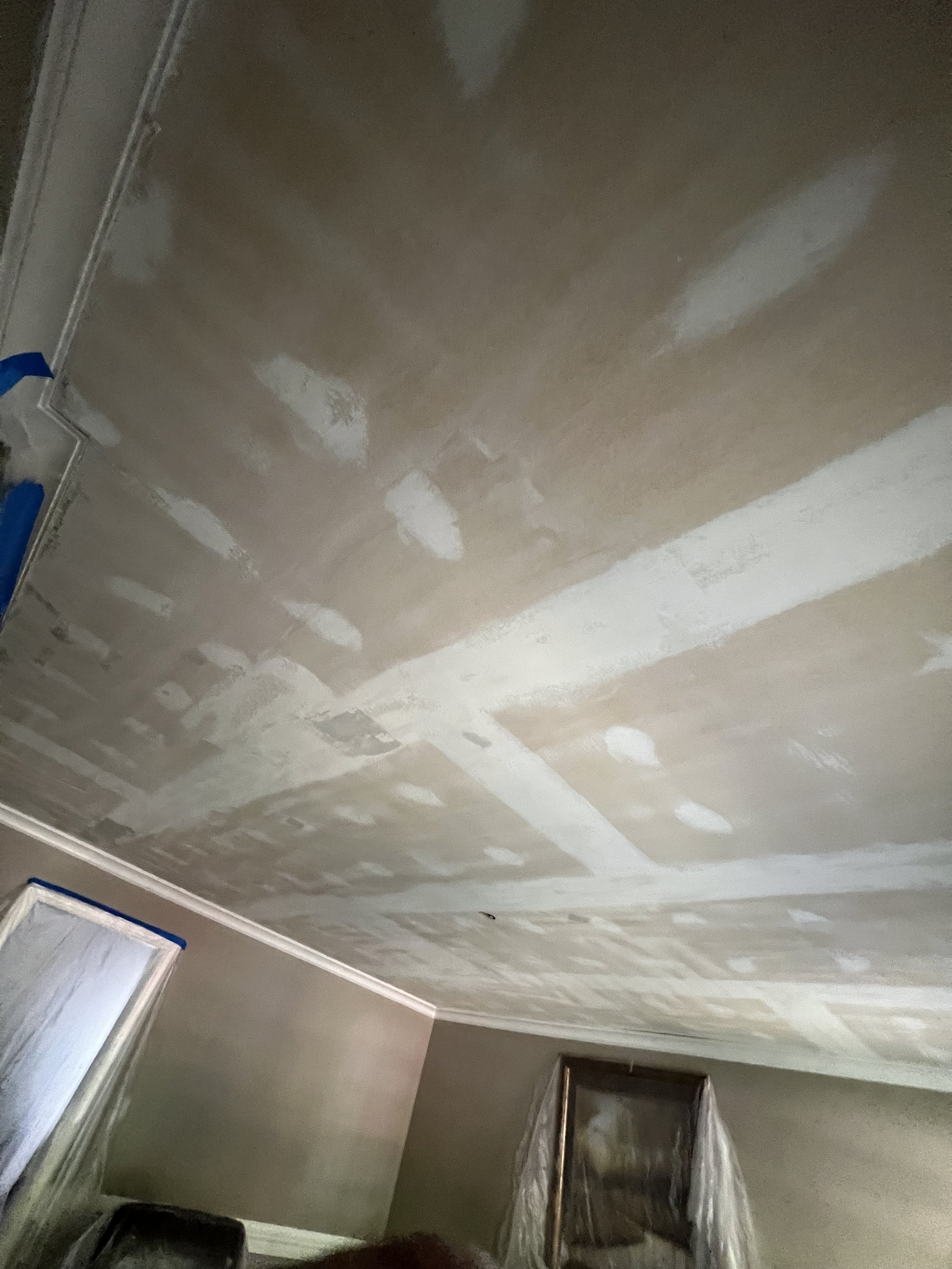 Unfinished drywall ceiling with patchy joint compound, in a room with plastic-covered window frames and a partially visible carpet.