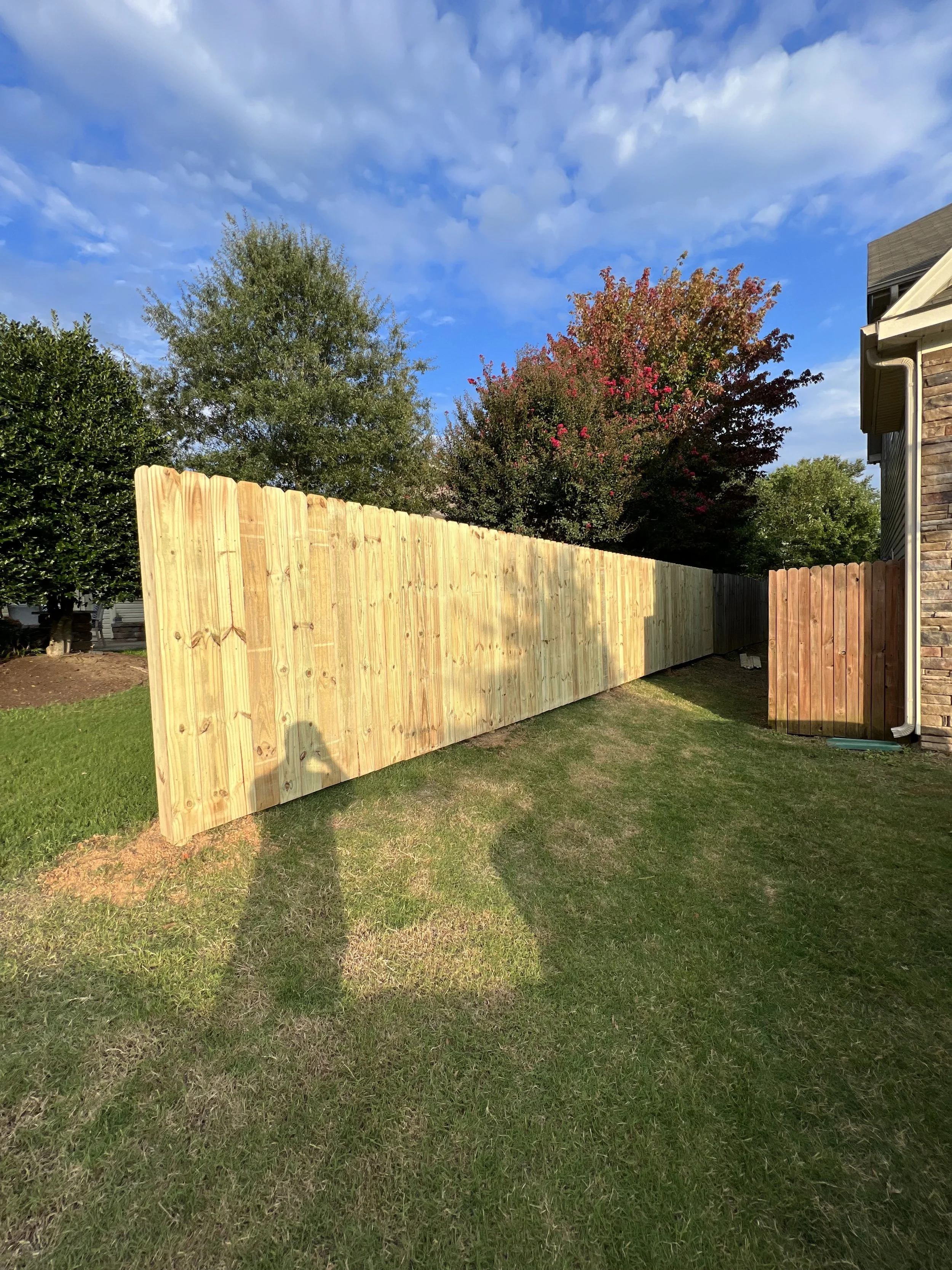 A wooden fence installed in a backyard, with grass and trees in the background and a partly cloudy sky.
