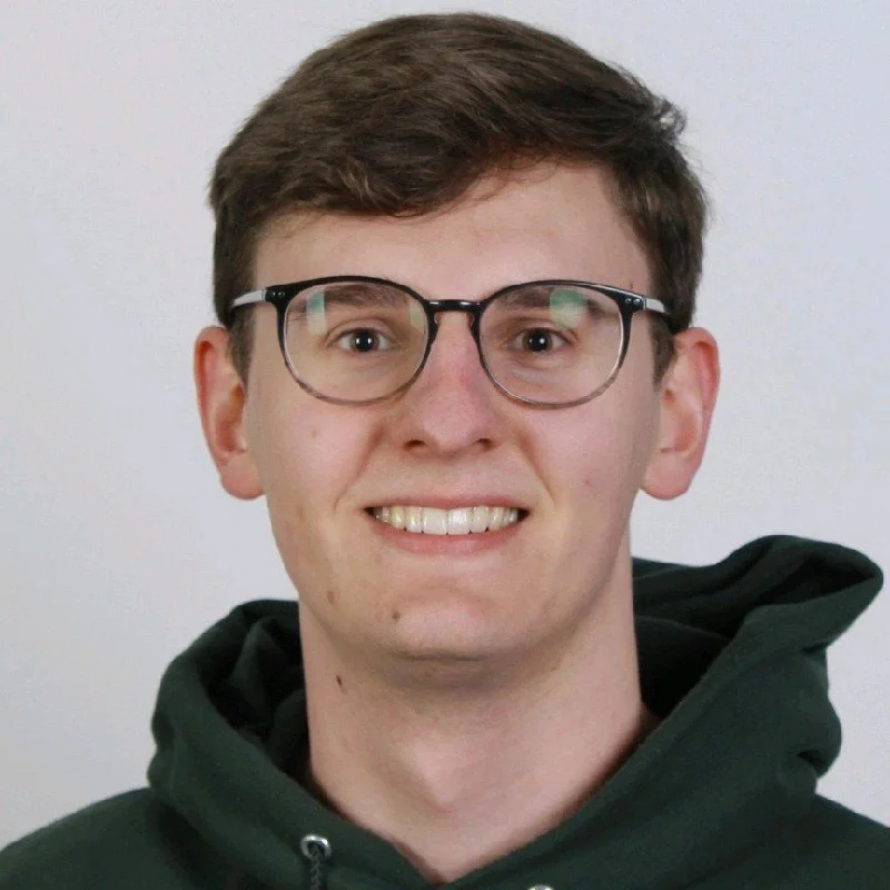 Young man with light skin, brown hair, wearing glasses and a dark green hoodie, smiling at the camera.