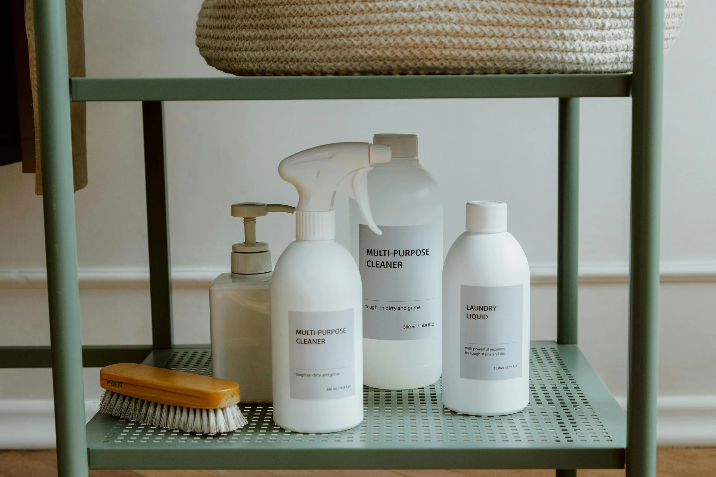 Cleaning supplies on a mint green metal shelves, including a soap dispenser, two spray bottles labeled 'Multi-Purpose Cleaner', a bottle of laundry liquid, a wooden scrub brush, and a container with a woven cover.