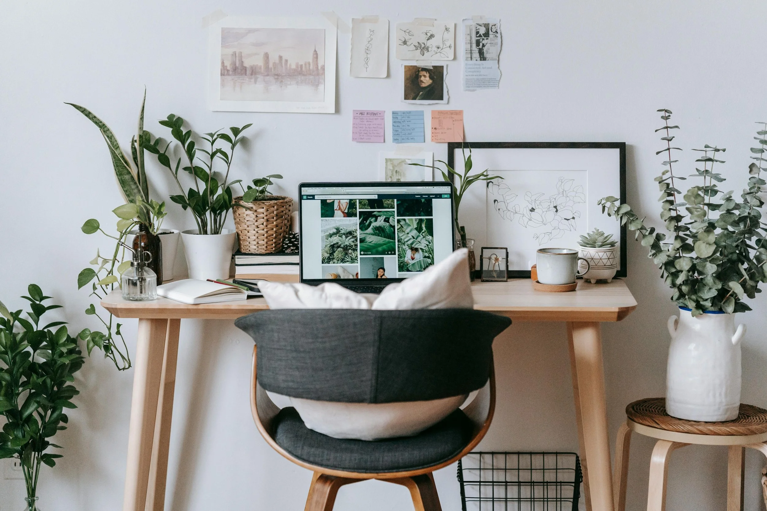 A cozy home office workspace with a wooden desk holding a laptop displaying a collage of nature photos, surrounded by potted plants, framed artwork, and stationery. A gray and white cushioned chair faces the desk, and a white vase with greenery is on a small wooden stool to the right.
