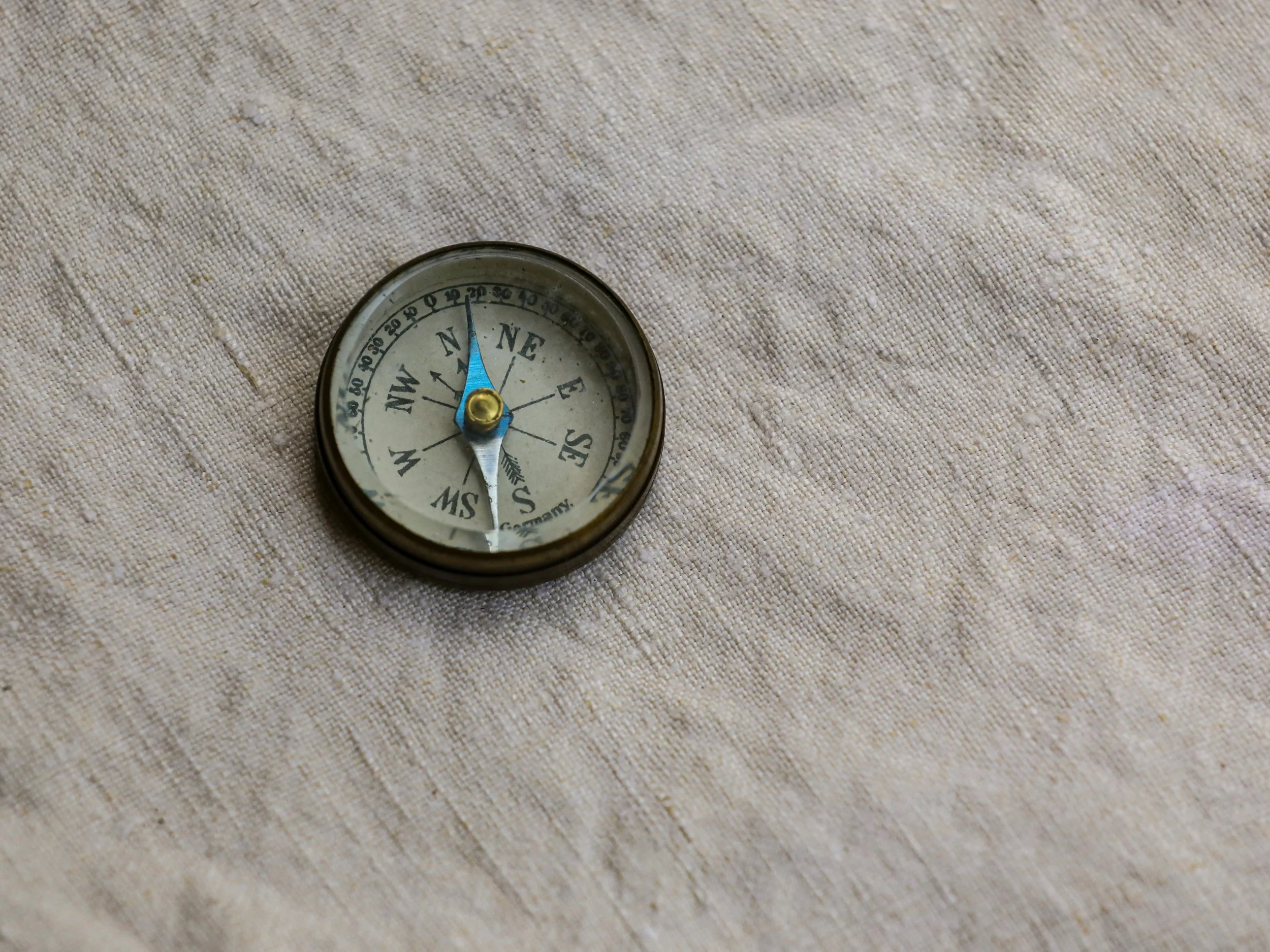 A vintage compass with a black rim and white face, showing north pointing slightly left of vertical, placed on a beige fabric surface.