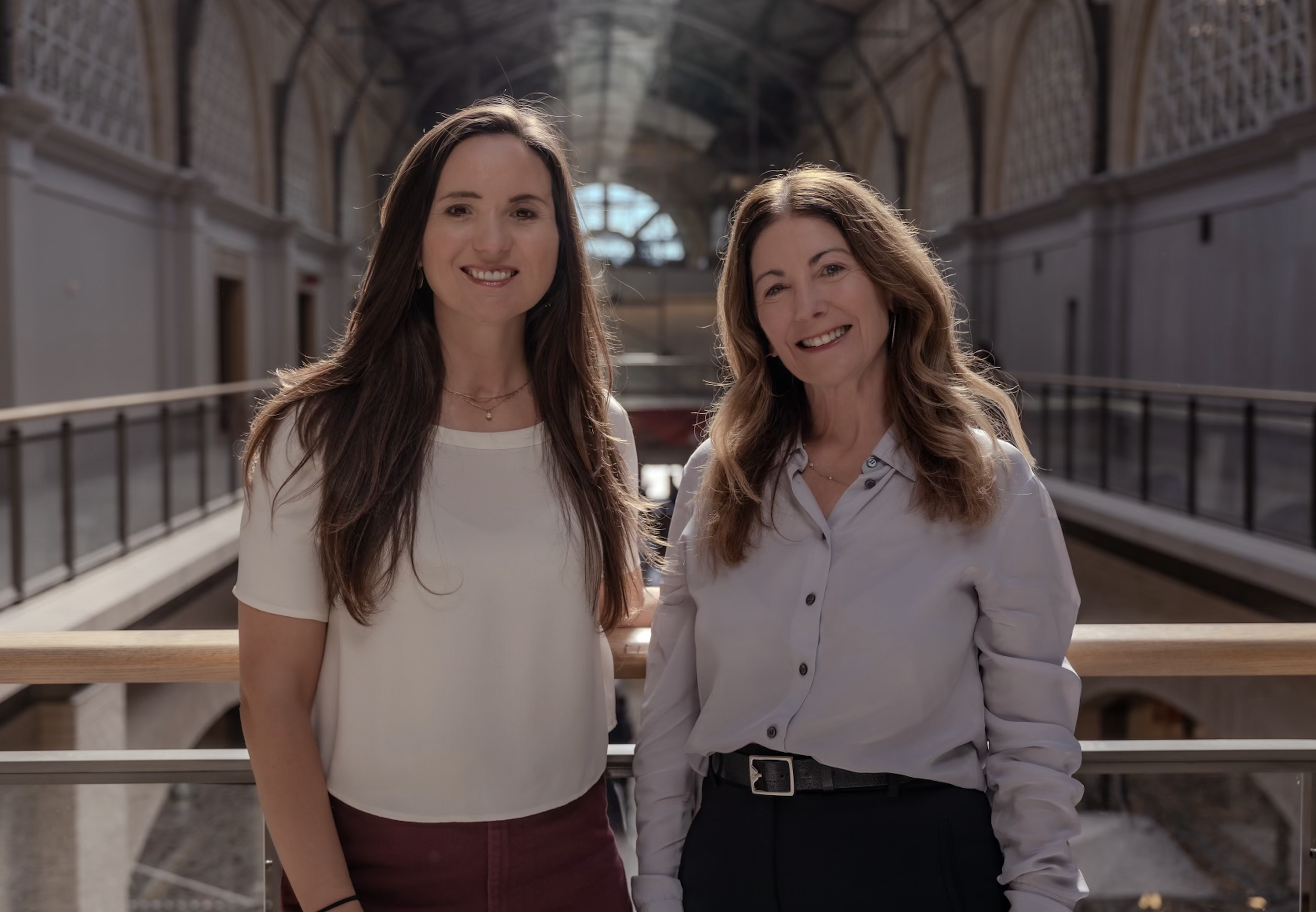 Two women smiling and standing in an indoor architectural setting with a high arched ceiling and windows in the background.